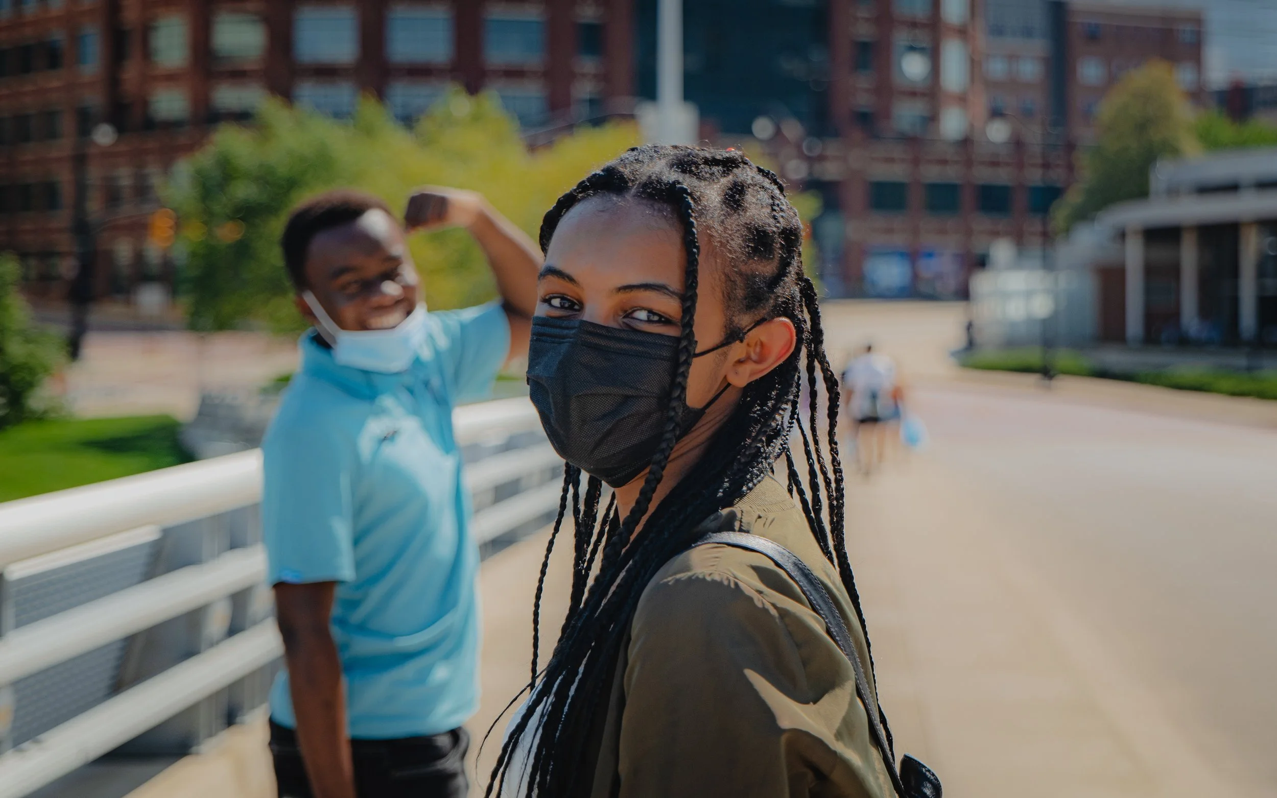 Two people outdoors wearing face masks, one smiling in the background, another with braided hair in the foreground. Urban setting with buildings and greenery.
