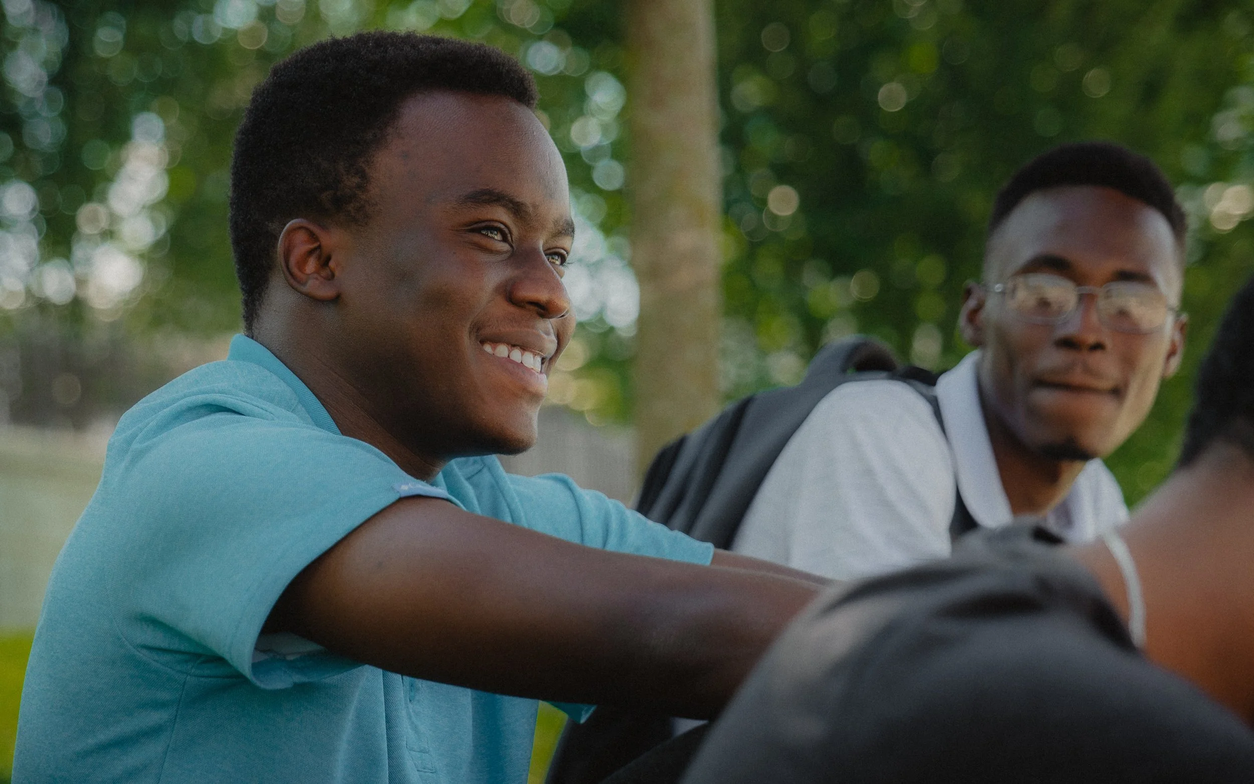 Two young men, one smiling in a blue shirt, the other wearing glasses, sitting outdoors.