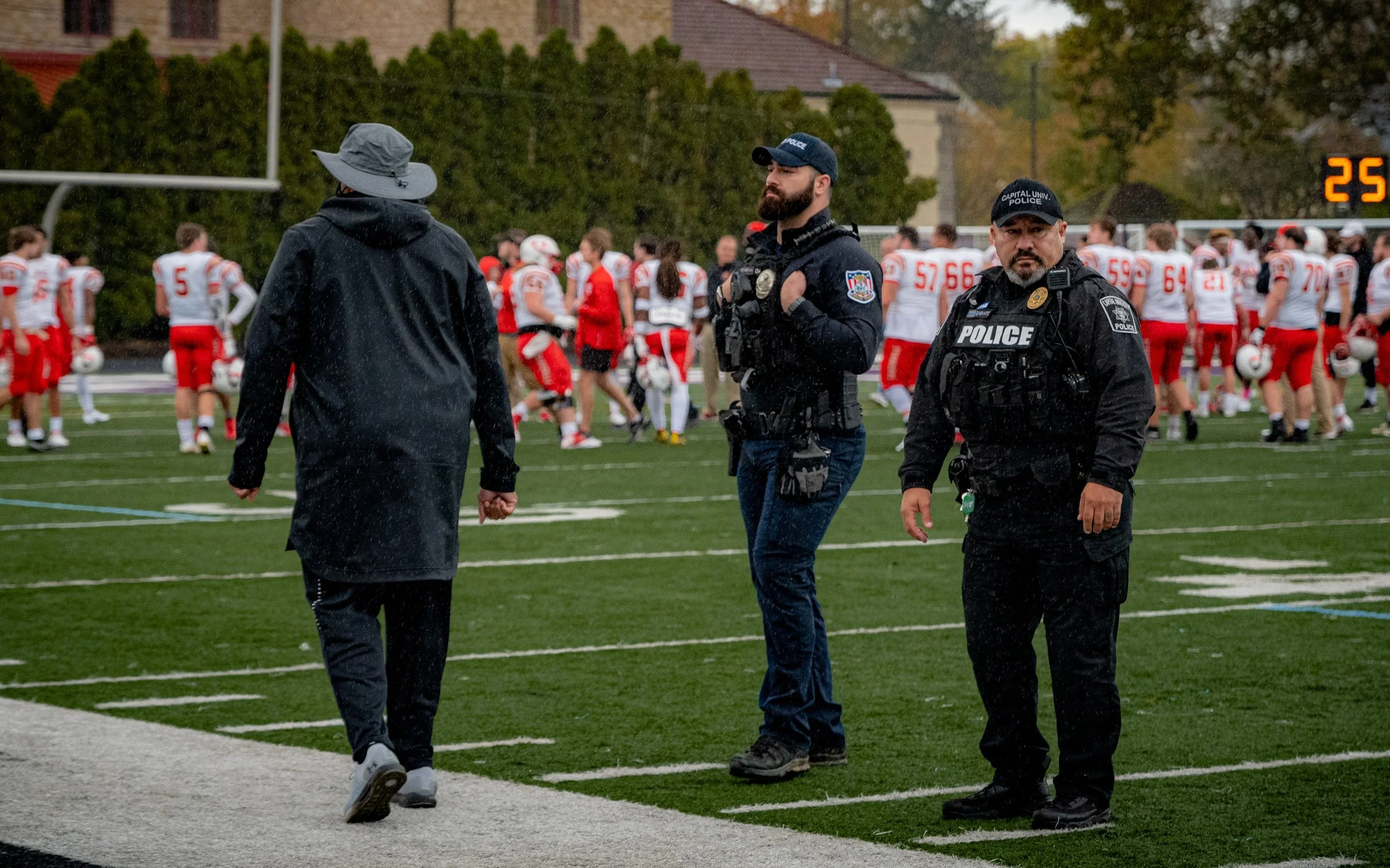 Police officers standing on the sidelines of a football field with a coach and players in uniforms in the background.