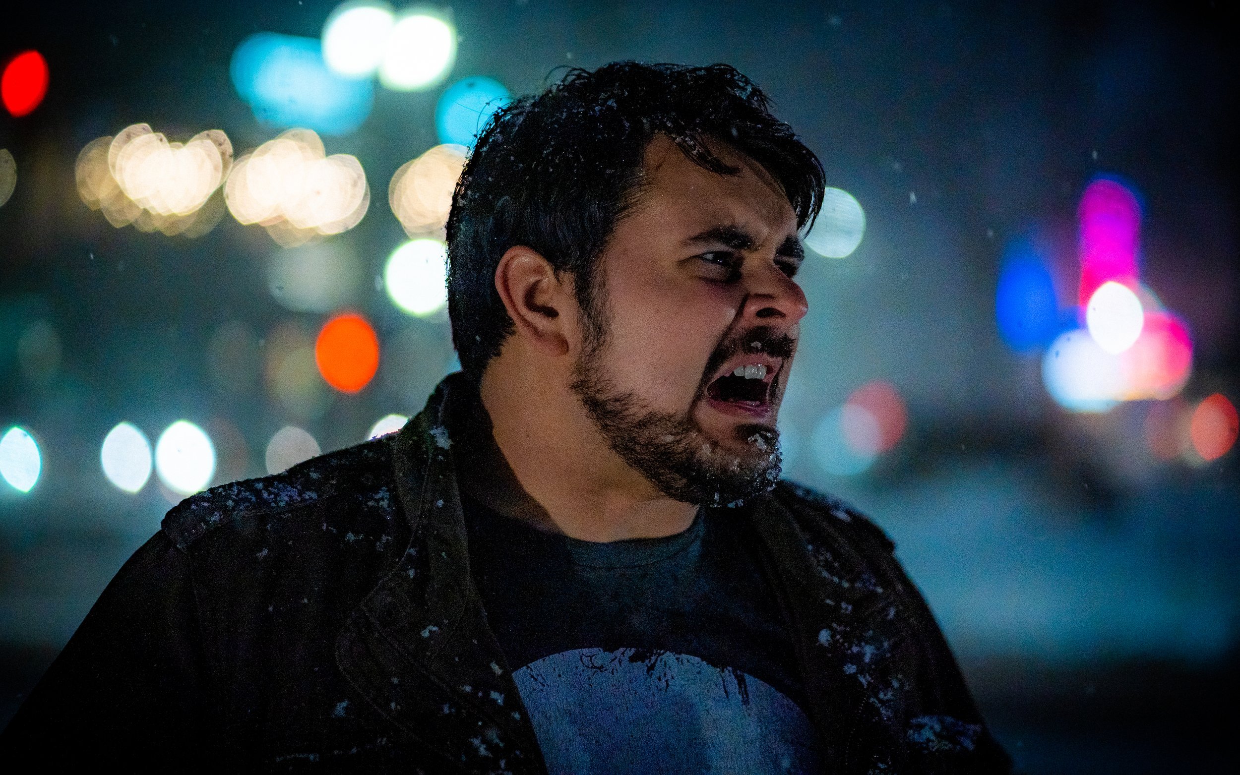 A man with a beard looking to the side, snowflakes on his shoulders and hair, expressing a strong emotion in a night setting with blurred colorful lights in the background.
