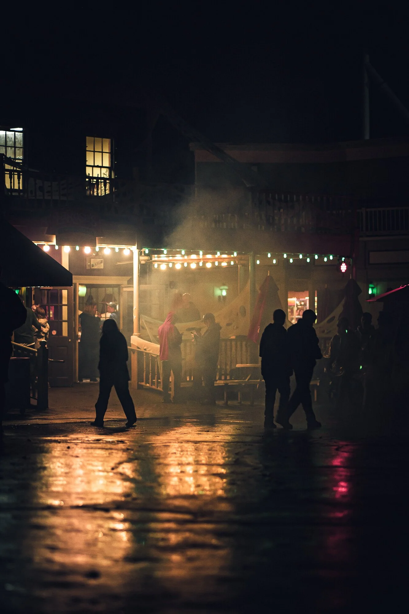 Nighttime scene with silhouettes of people walking on a wet street outside a lit-up building. Colored lights and a foggy atmosphere create a moody ambiance.
