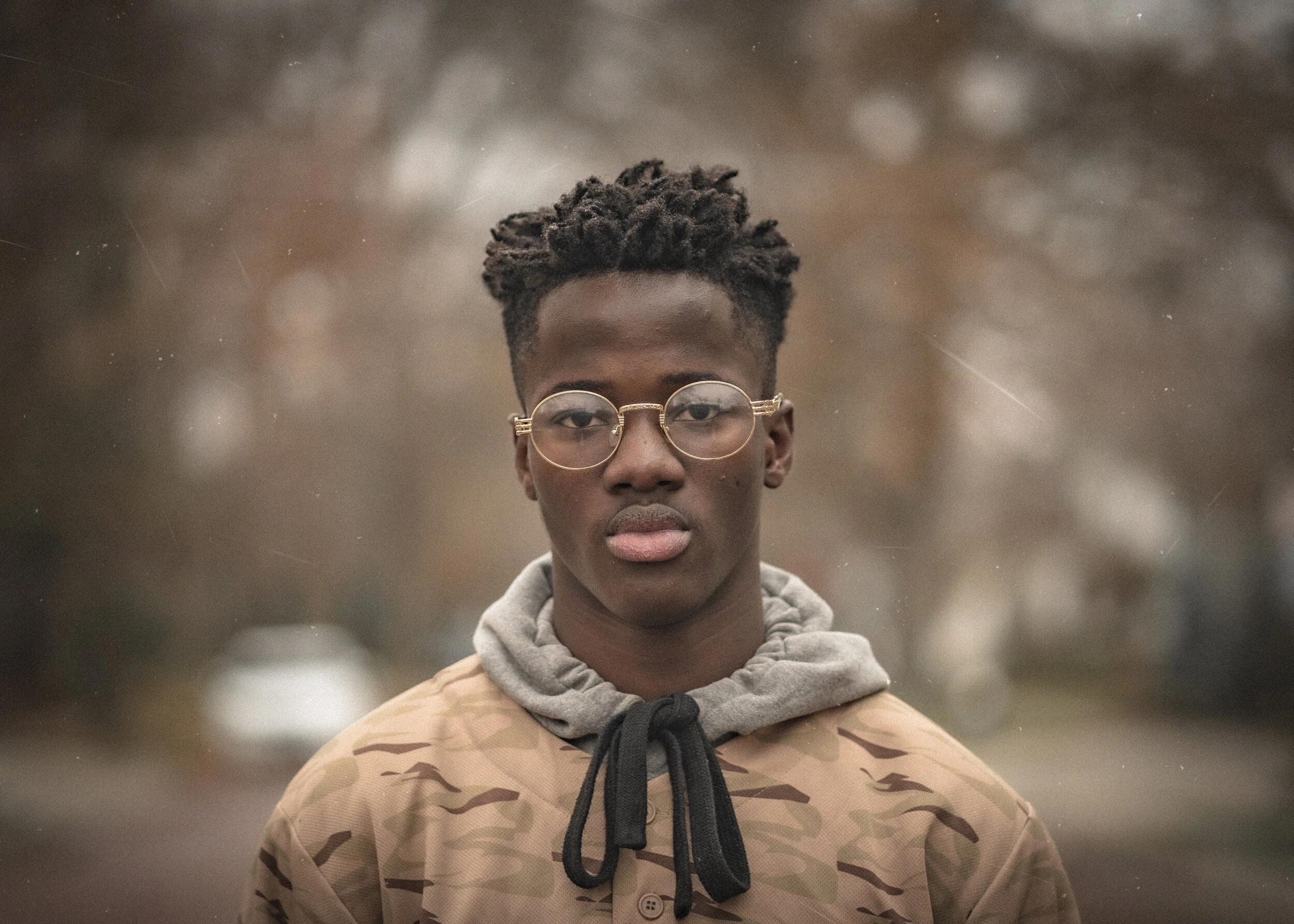Young man wearing glasses and a camouflage jacket standing outdoors with a blurred background.