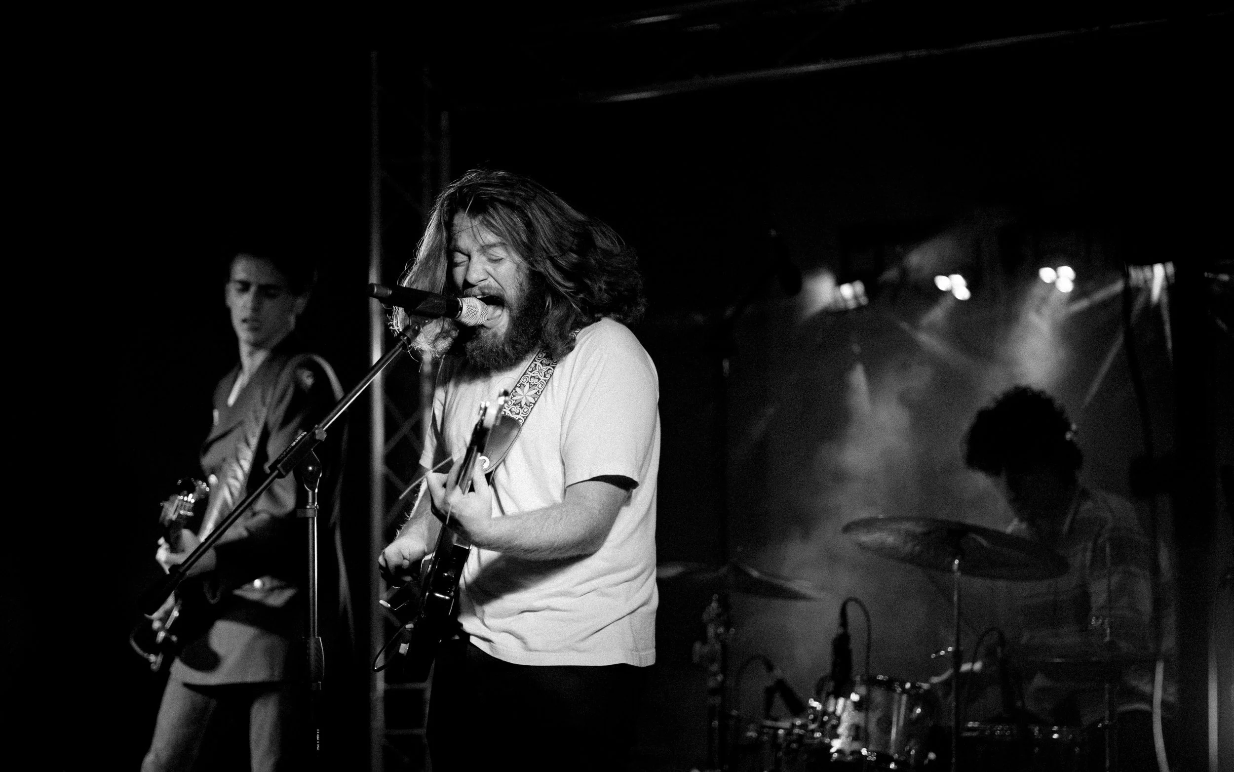 Black and white photo of a band performing on stage, featuring a guitarist singing into a microphone with other musicians in the background playing guitar and drums.