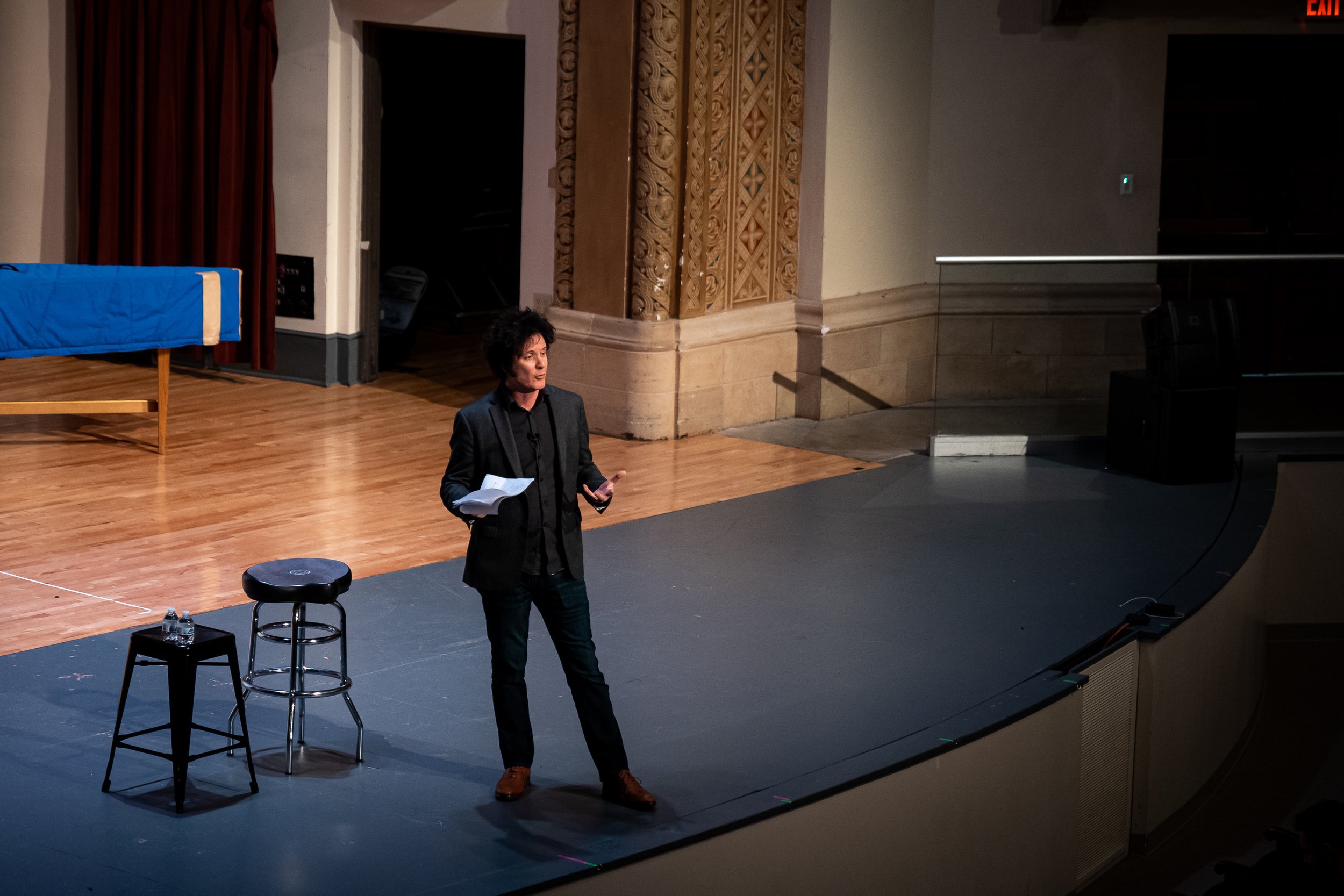 Person giving a lecture on a stage with a microphone and papers in hand, next to two stools.