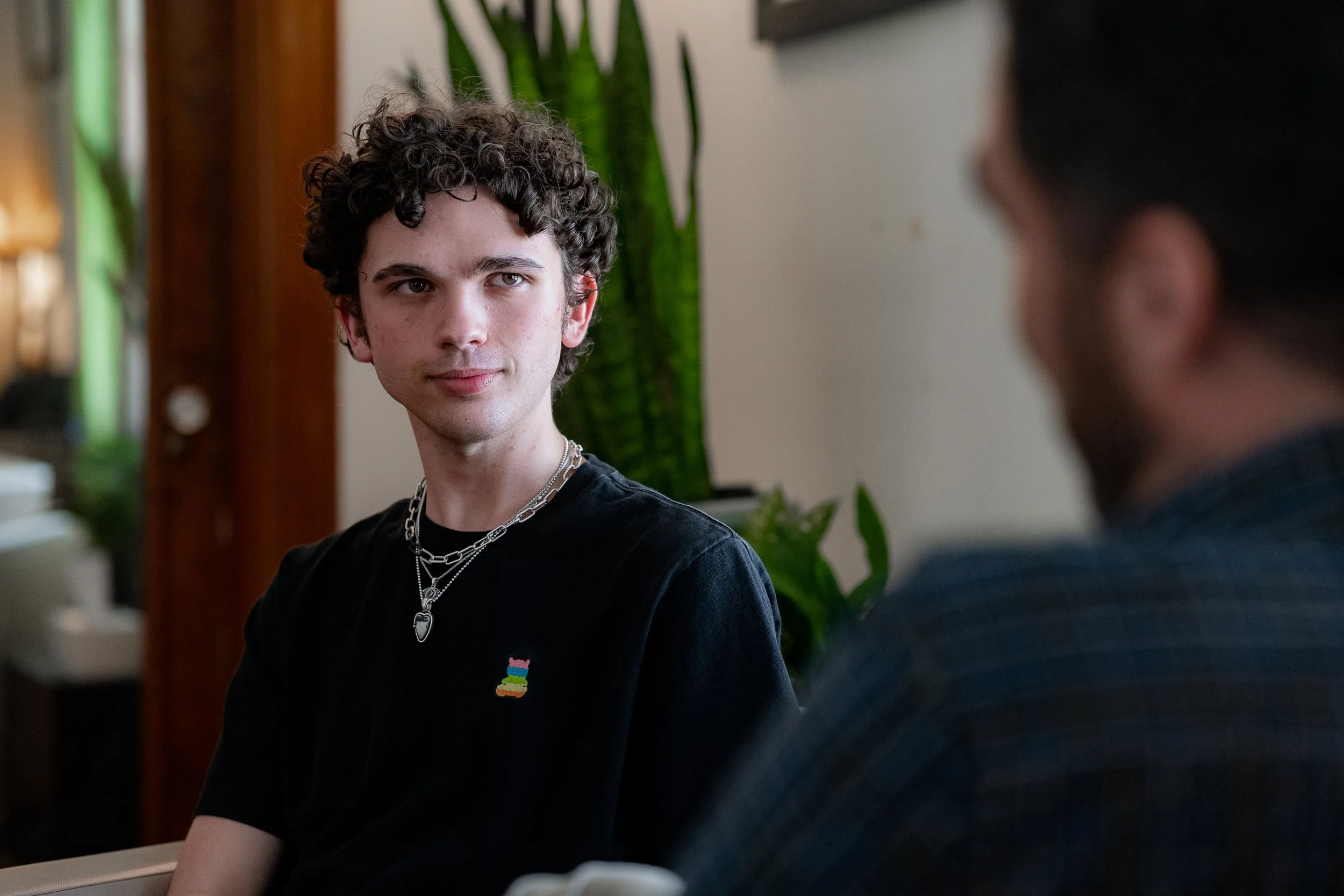 Two people having a conversation indoors, with one person in focus wearing a black T-shirt and necklaces, and plants in the background.