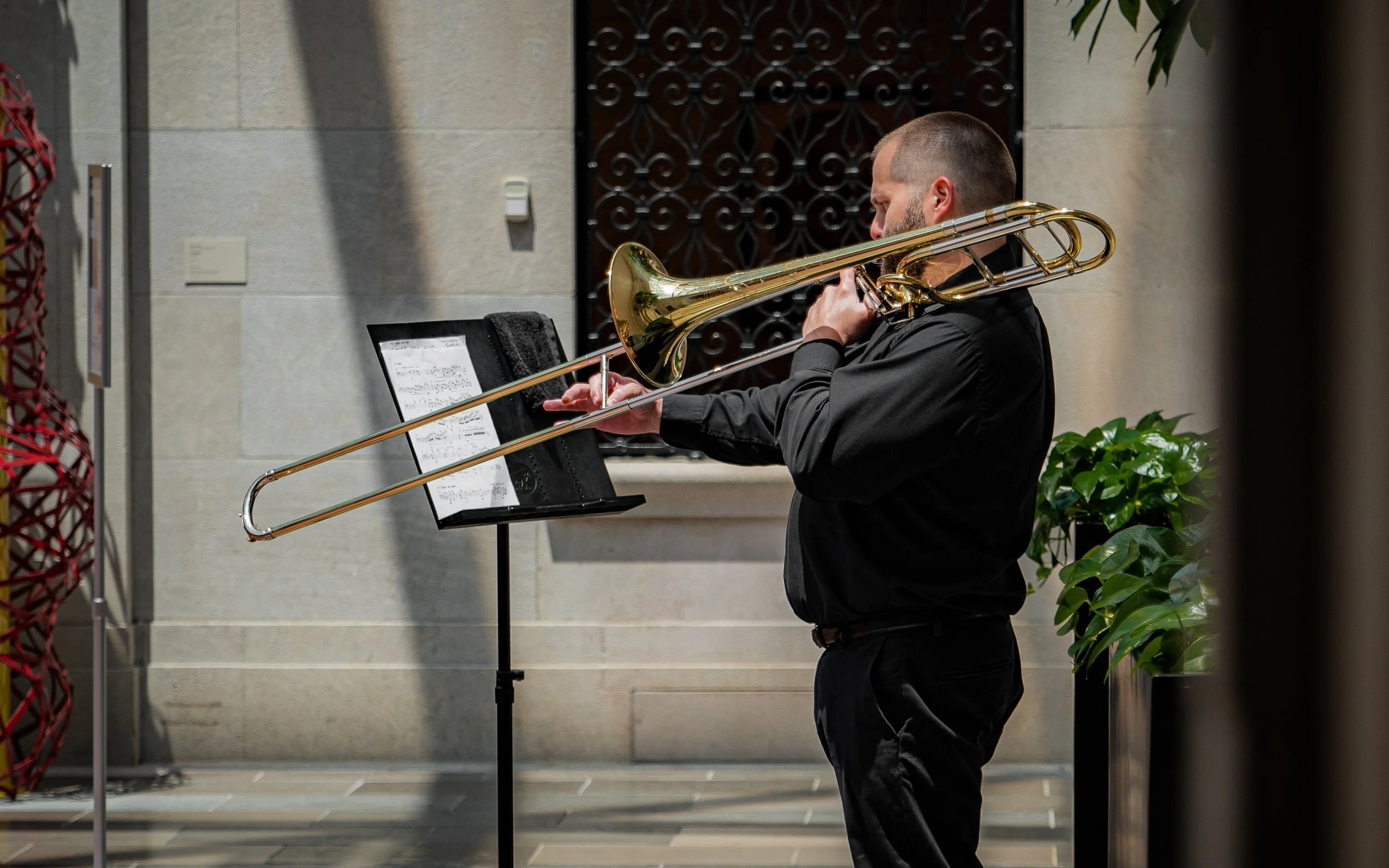 Man playing the trombone in an indoor setting with sheet music on a stand; plants and decorative elements in background.