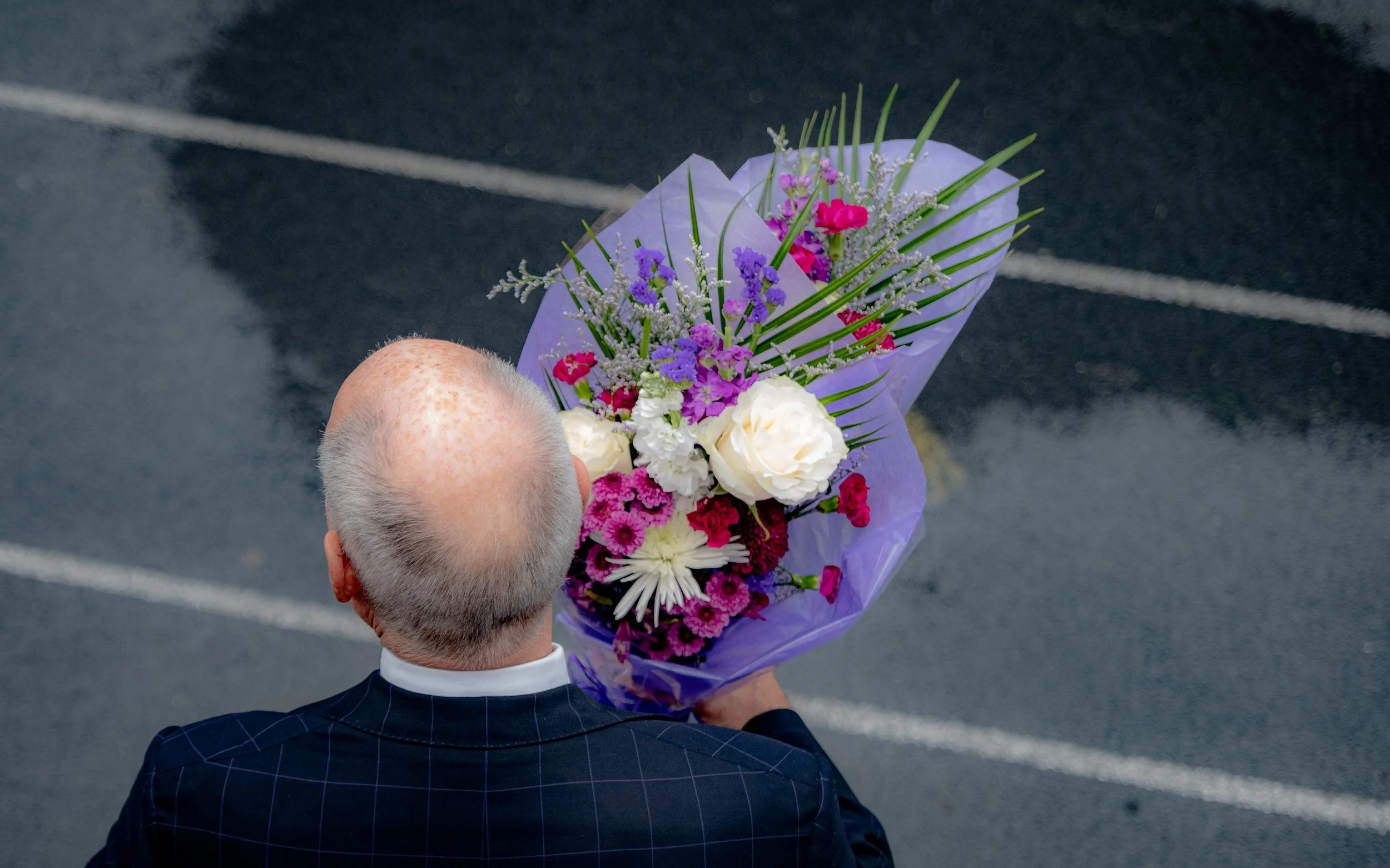 Bald man holding a bouquet of flowers wrapped in purple paper, viewed from behind on a wet pavement.