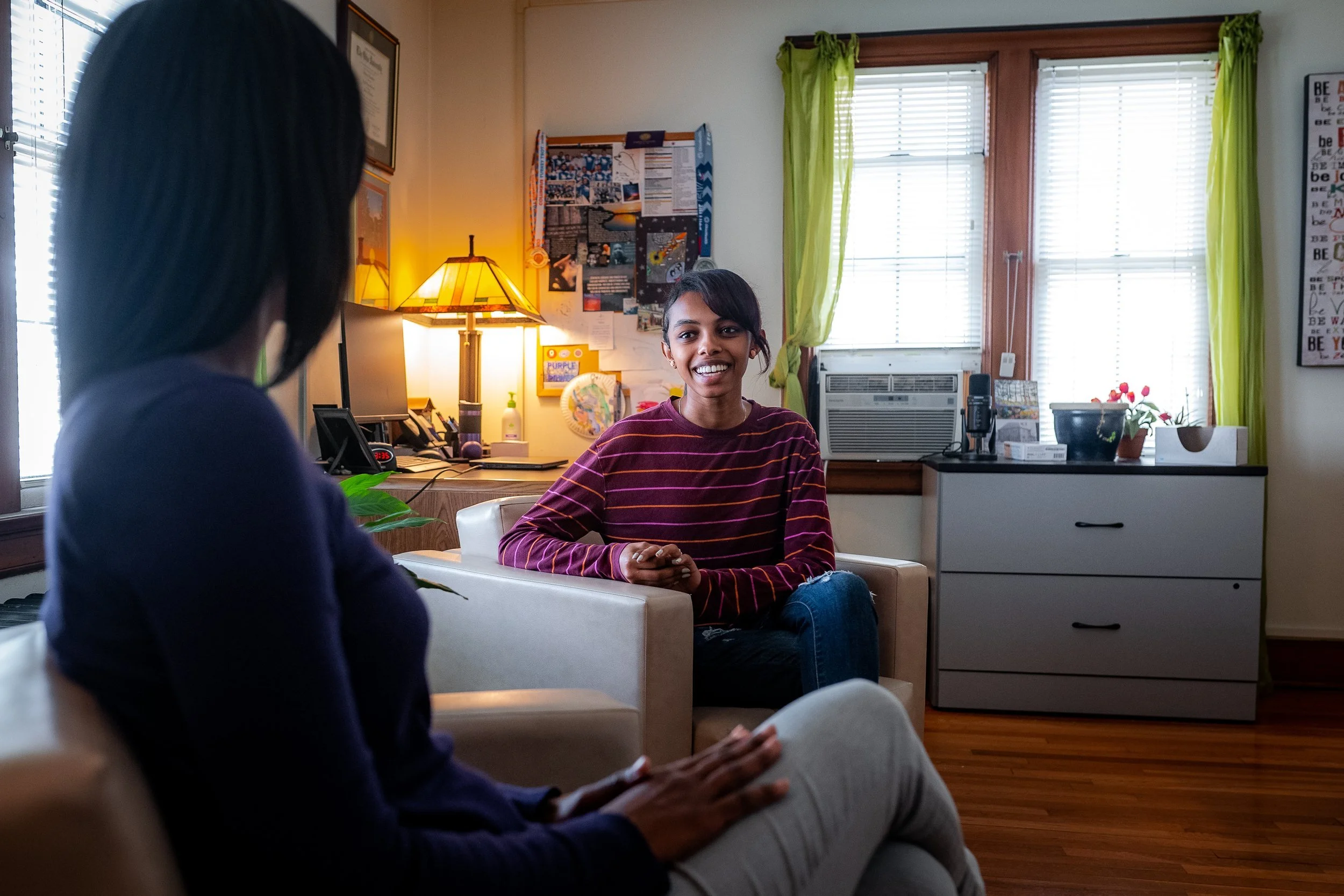 Two people sitting and talking in a cozy office with a lamp, desk, and bulletin board in the background. One person is smiling and wearing a striped sweater, while the other is seen from behind.