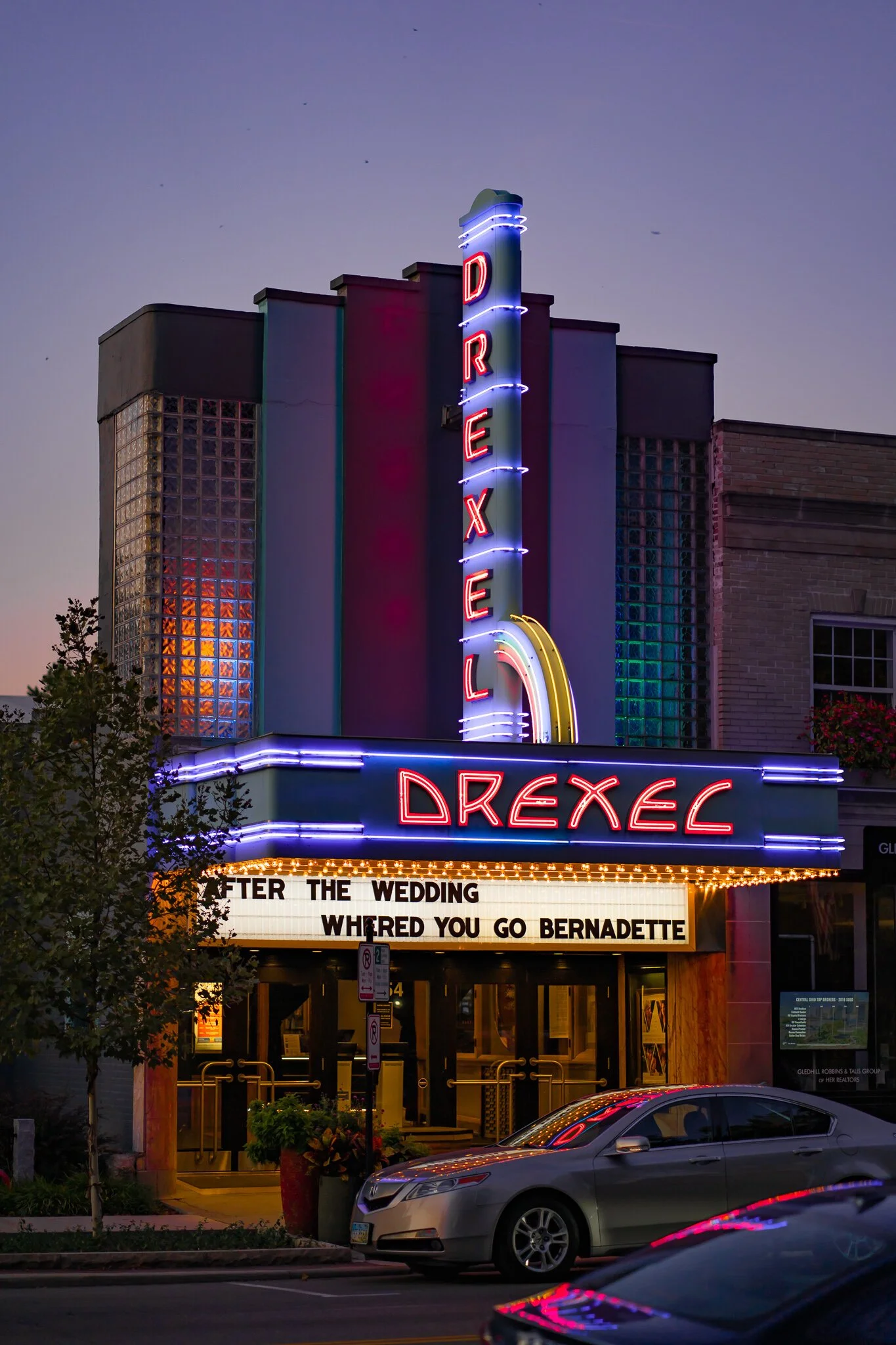 Exterior of Drexel Theatre at dusk with neon lights and marquee displaying movie titles.