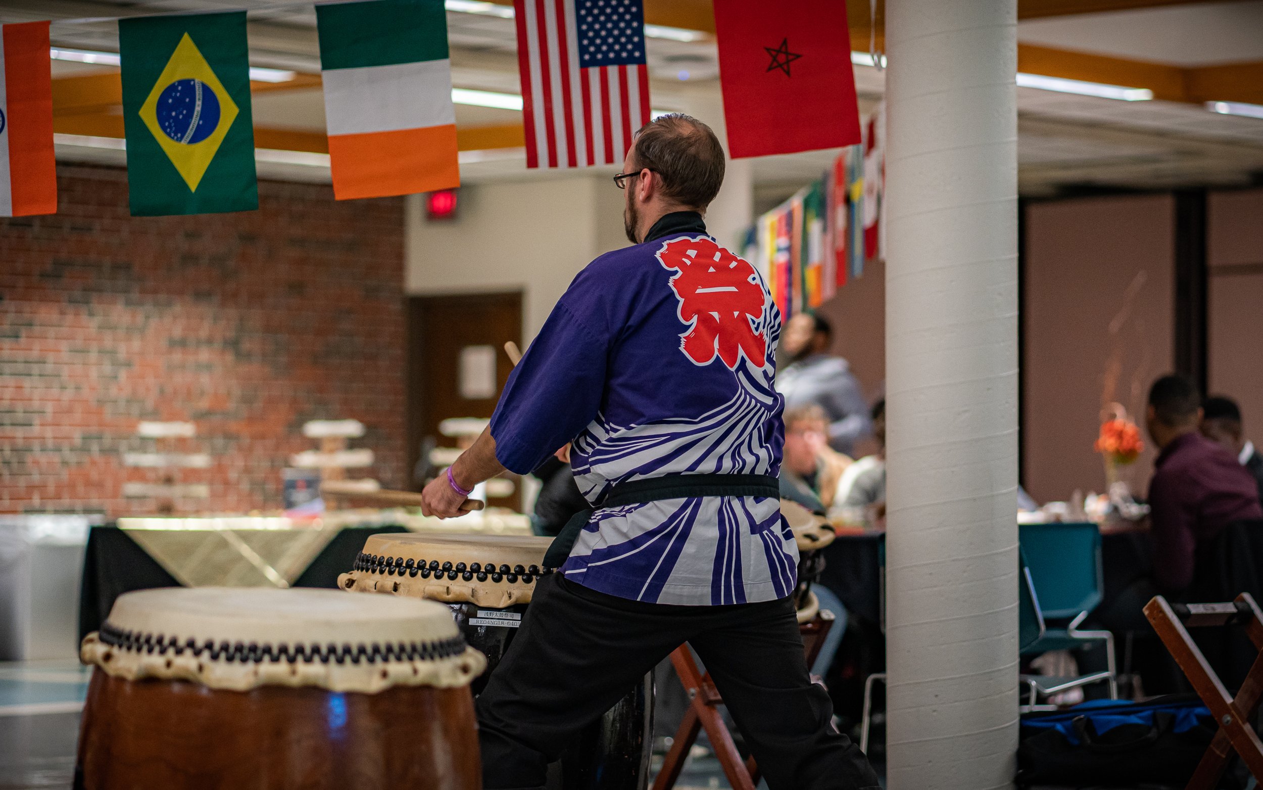 A person playing a traditional drum wearing a colorful robe with flags hanging overhead, including Brazilian, Irish, American, and others.