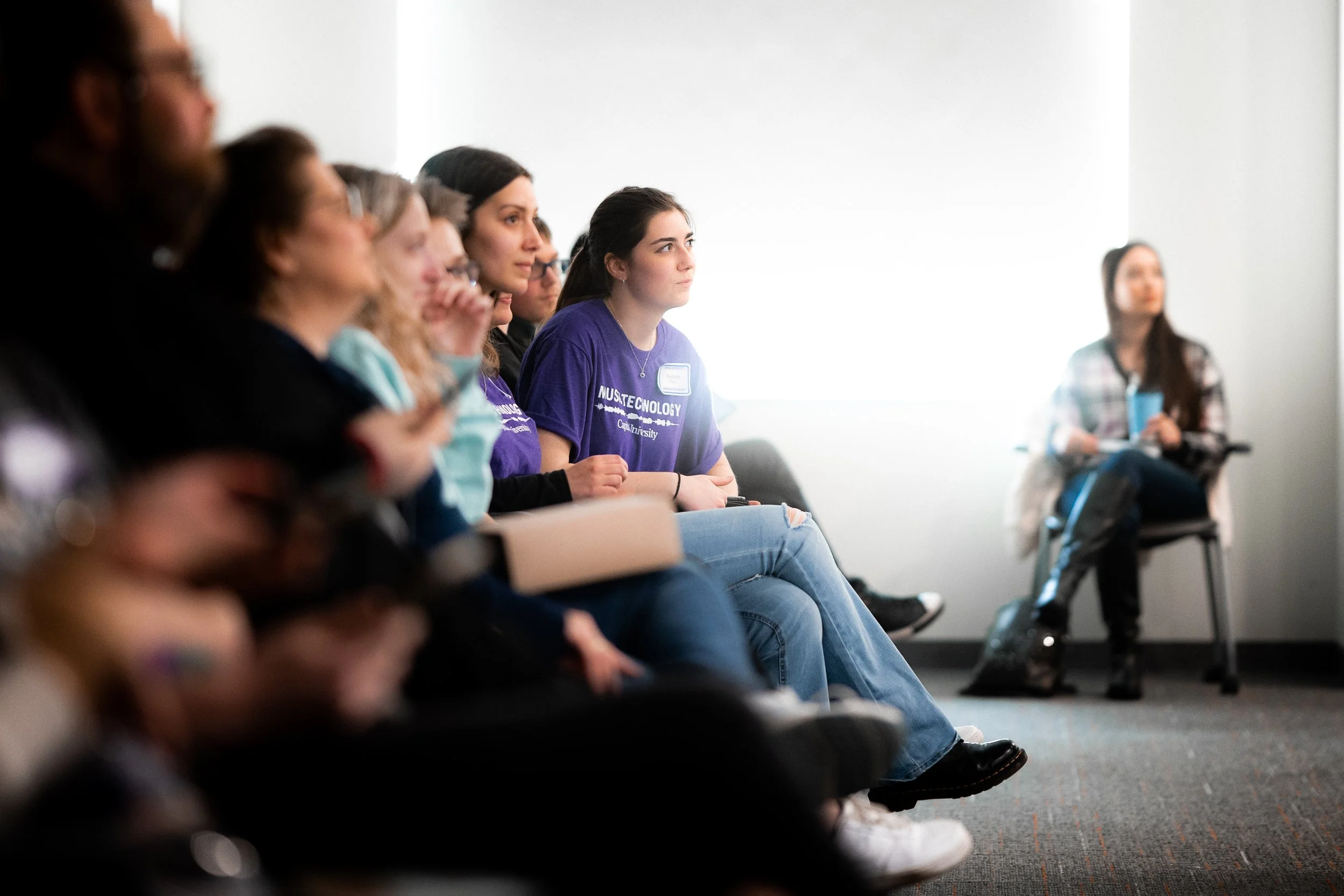 A group of people sitting in a room, facing towards the right side. Some are on chairs, attentive, with a woman in a purple shirt in focus. The setting appears educational or seminar-like.