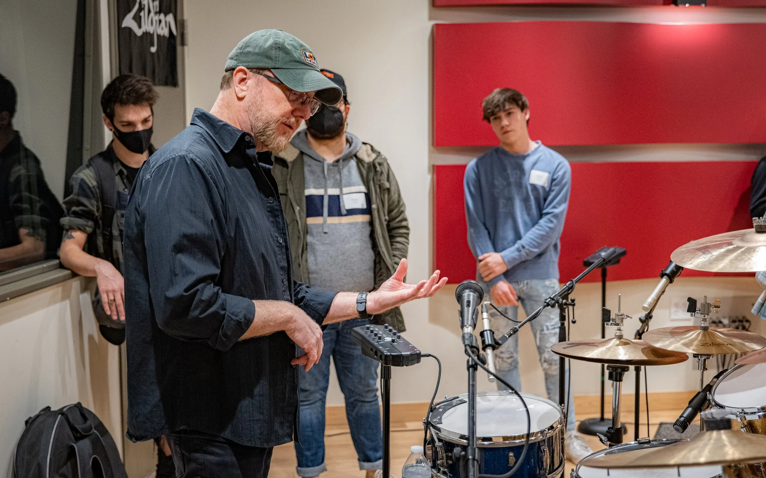 A man wearing a hat and glasses is speaking to a group of people in a recording studio. He is standing near a drum set with microphones. The people around him are listening attentively, some wearing masks. The studio has red acoustic panels on the wa