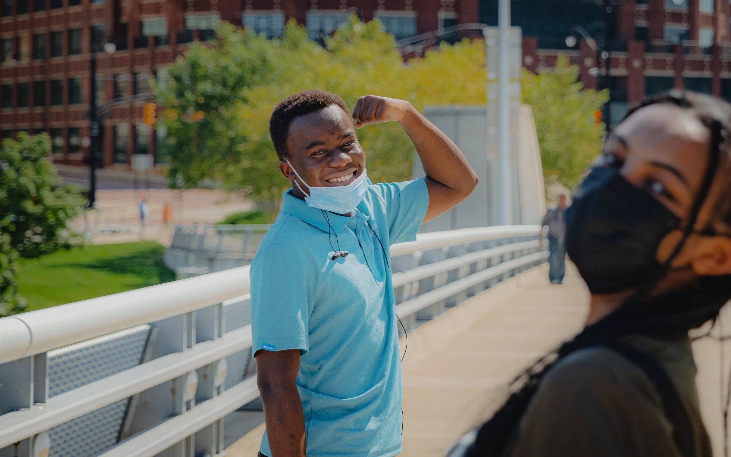 Two people on a bridge wearing face masks; one is flexing his arm and smiling, the other is slightly blurred in the foreground.