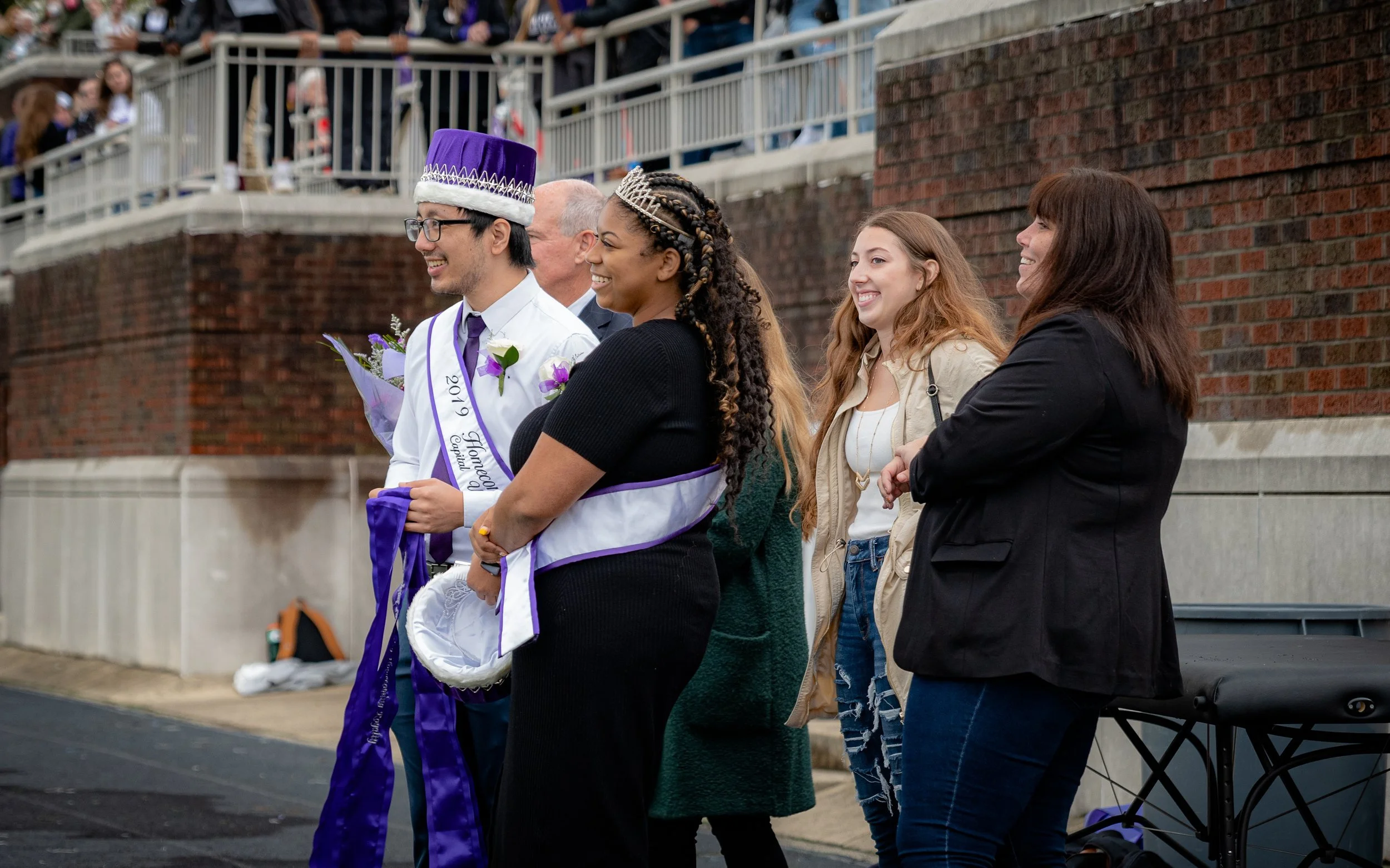 Group at outdoor event with two people wearing sashes and crowns, smiling with onlookers in the background.
