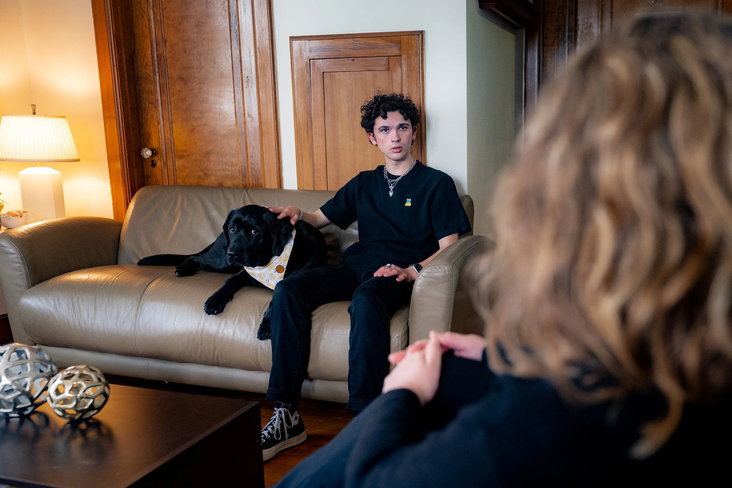 A person sitting on a couch with a black dog wearing a bandana, in a cozy room with wooden doors and lamp beside them.