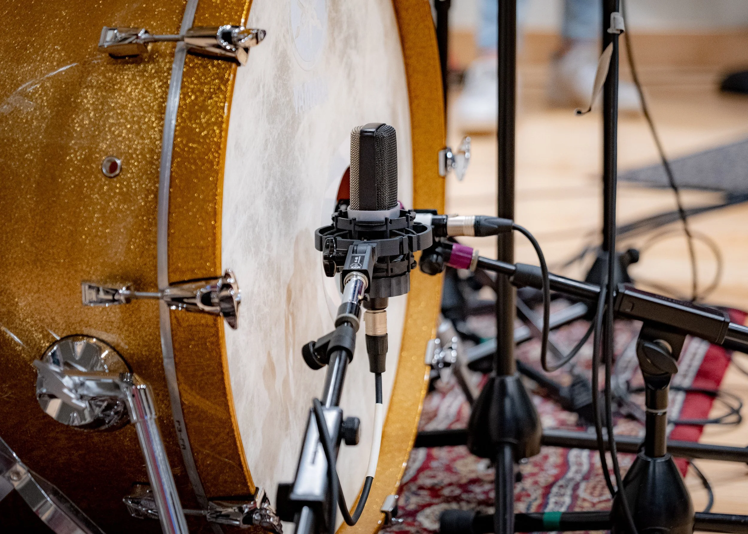 Close-up of a golden bass drum with a microphone placed in front of it, capturing sound. Multiple cables and mic stands are visible around the drum on a rug.