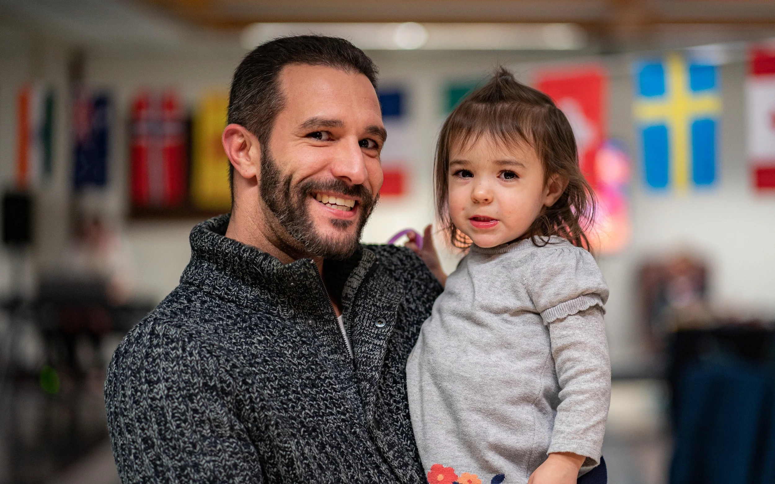 A smiling man with a beard holding a young child in a room decorated with international flags.