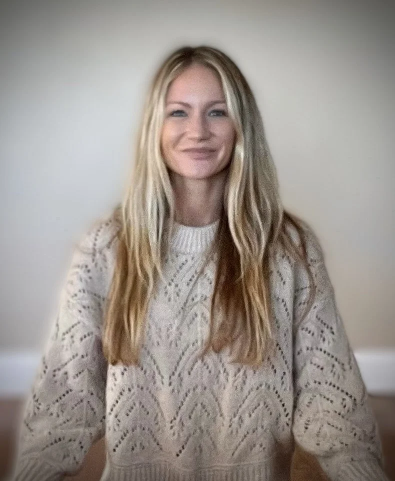 A young woman with long blond hair, wearing a cream knit sweater, smiling at the camera against a plain light-colored wall.