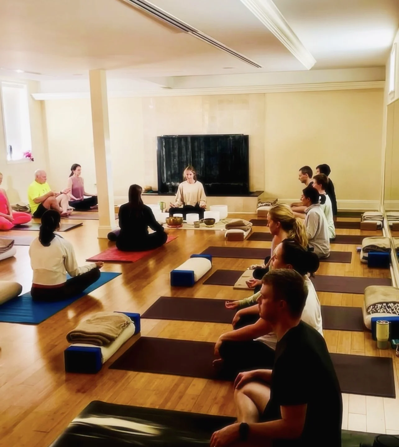 Group of people participating in a meditation or yoga class in a bright, spacious studio with wooden floors and a large television at the front.