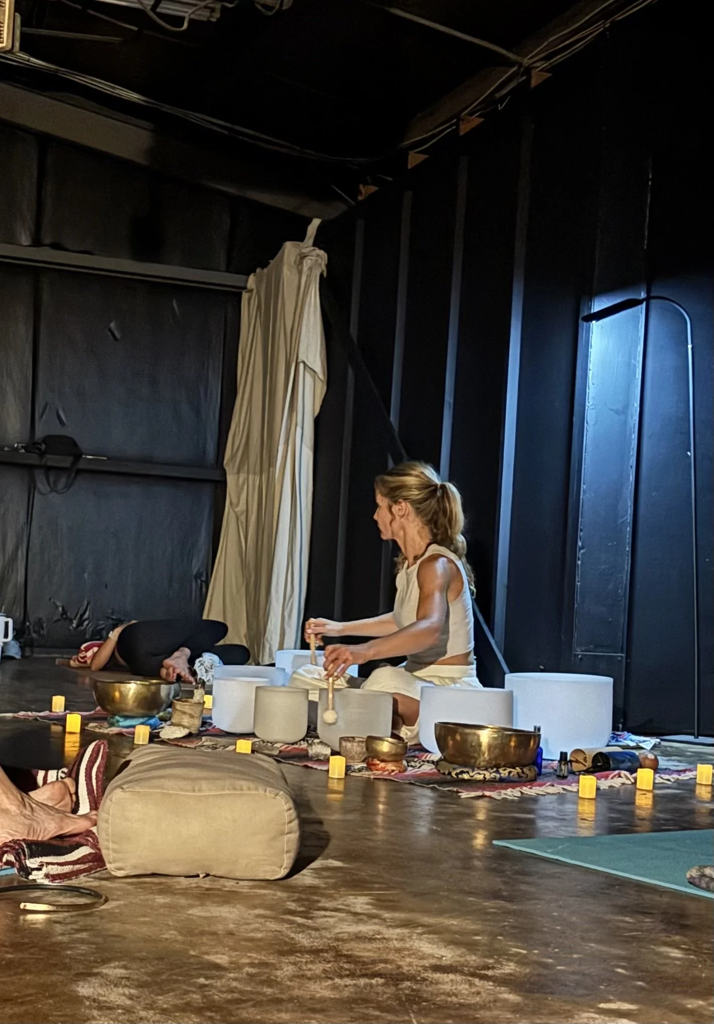 A woman in a white dress is sitting on the floor playing singing bowls, with other people lying down on the floor, some with their heads resting on cushions. The environment looks like a meditation or sound healing session with candles and soft lighting.