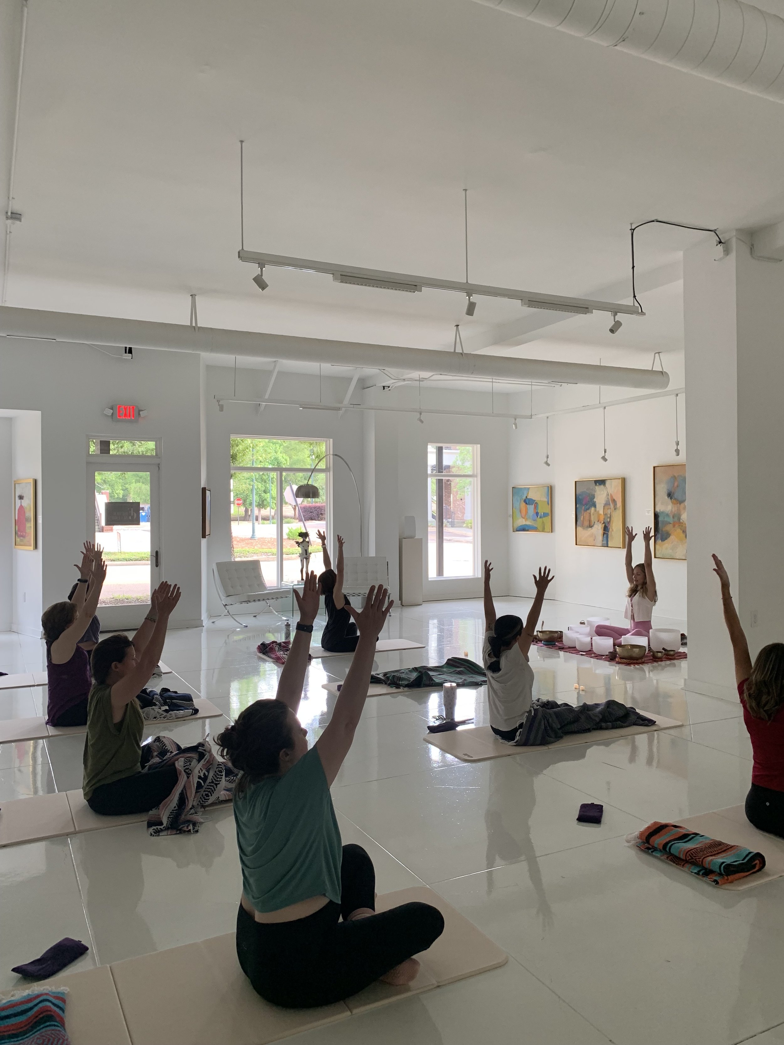 A group of people participating in a yoga or meditation class in a bright, modern studio with white walls, large windows, and colorful artwork. Participants are seated on mats with their arms raised, led by an instructor sitting on a raised platform.