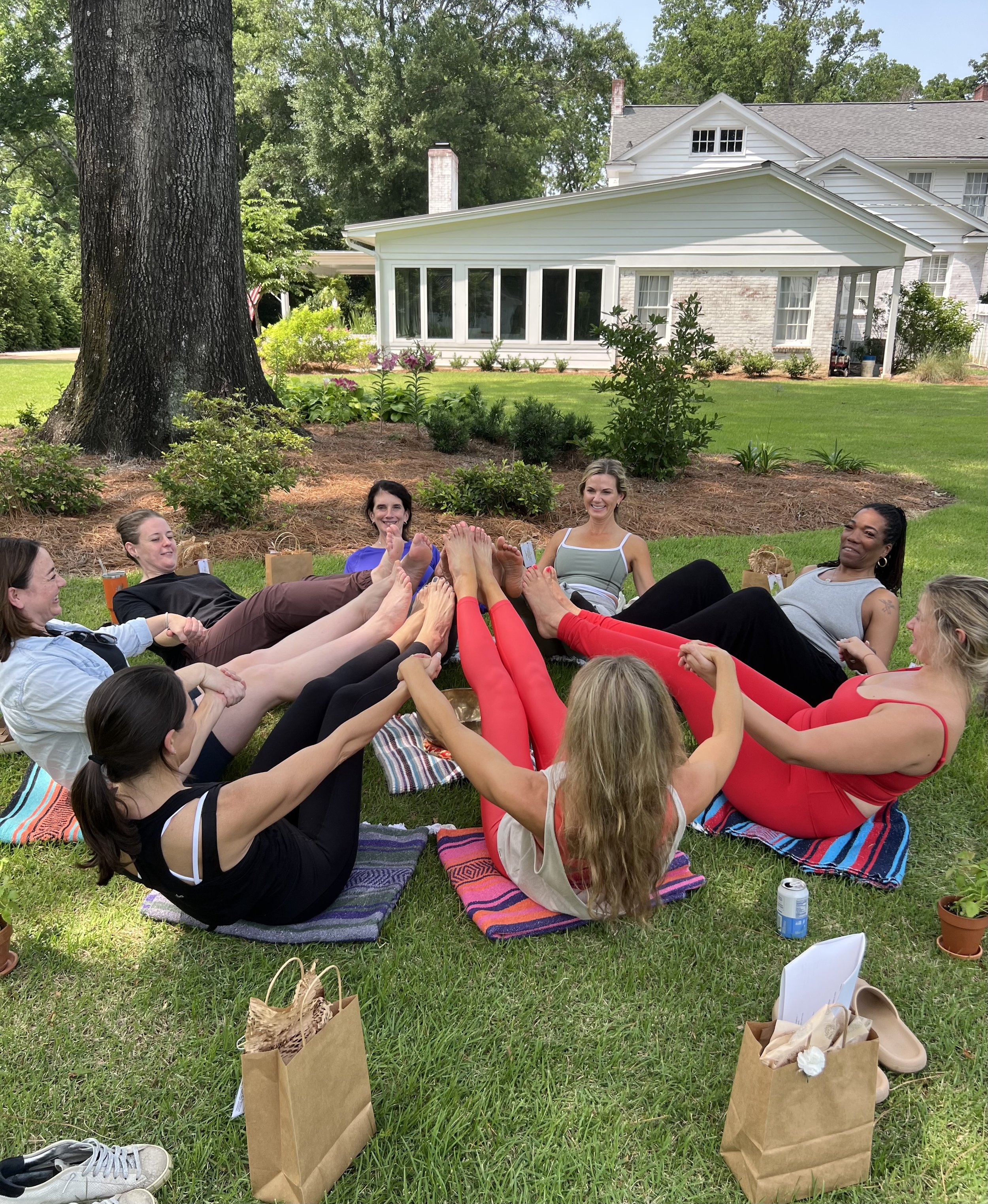 Group of women sitting outdoors on a grassy area, engaged in a circle activity, with shopping bags in front of them, a large tree, and a white house with a porch and screened-in area background.
