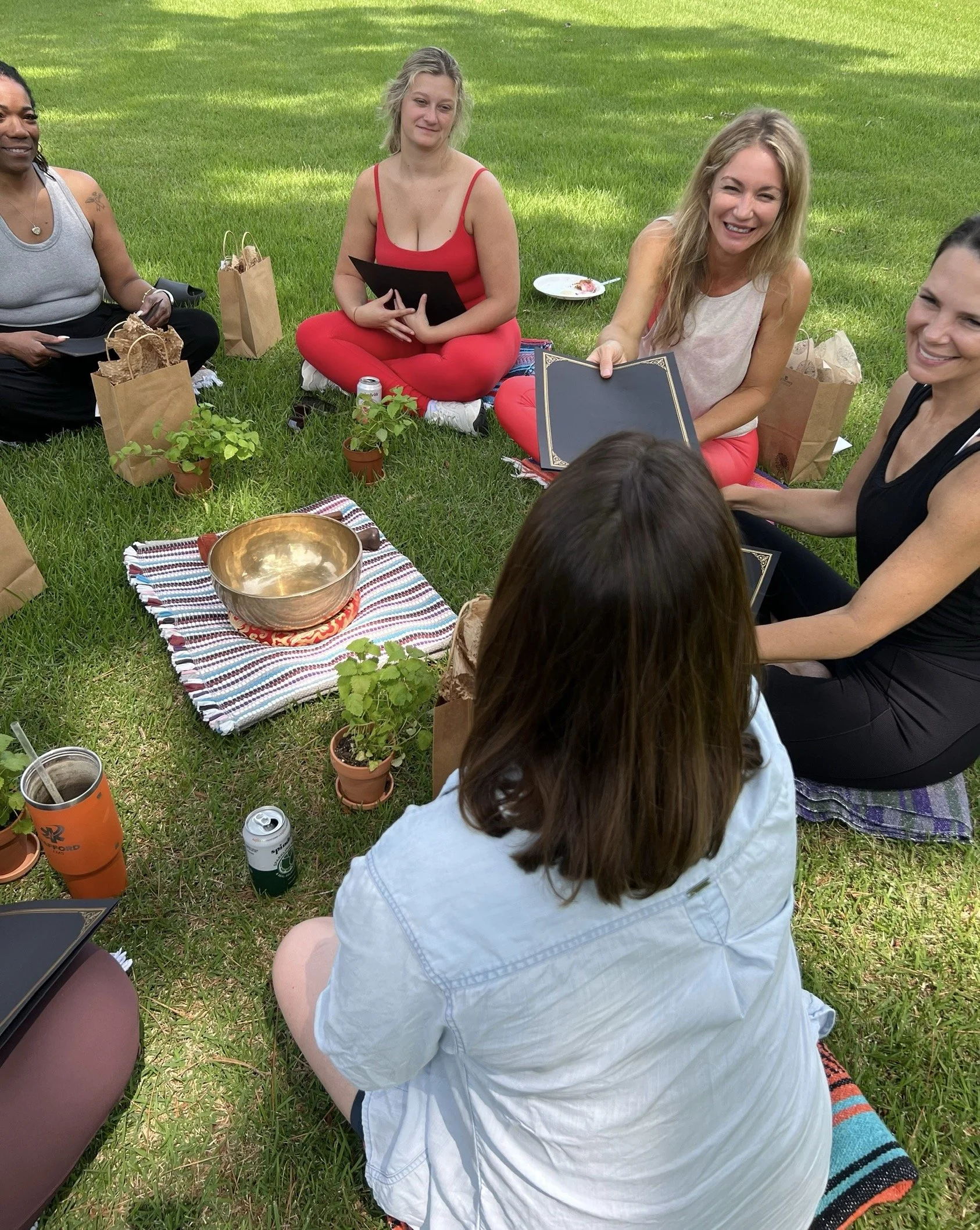 A group of women sitting on the grass during a picnic, smiling and holding black photo albums. There are potted plants, paper bags, food plates, and drinks around them.