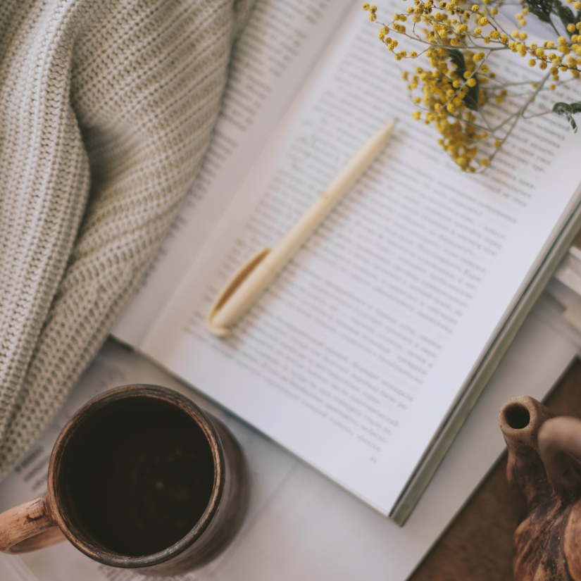 Open book with a cream-colored pen on top, a mug of coffee below, yellow flowers on the upper right, and part of a knitted blanket on the left.