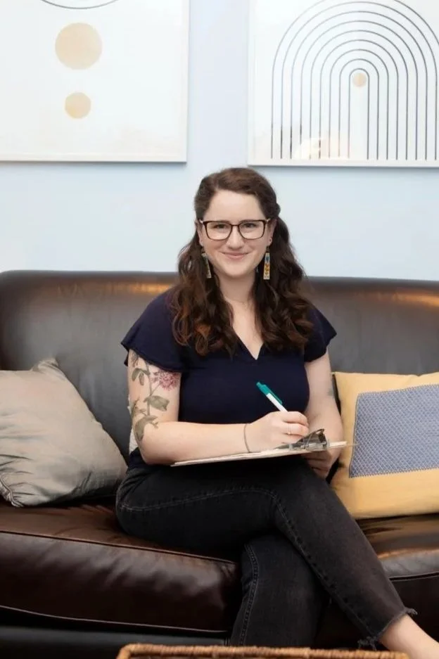 Person with long hair, glasses, and tattoos sitting on a leather couch, holding a clipboard and pen, smiling at the camera, with abstract wall art behind her.