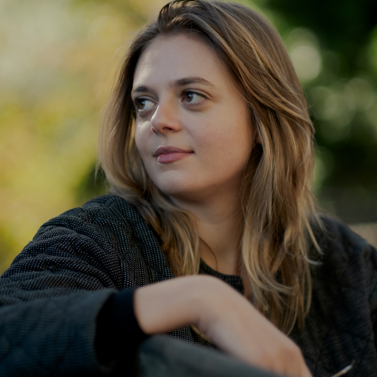 A young woman with light brown, wavy hair, wearing a black jacket, is sitting outdoors with a blurred green background, looking to her left with a slight smile.