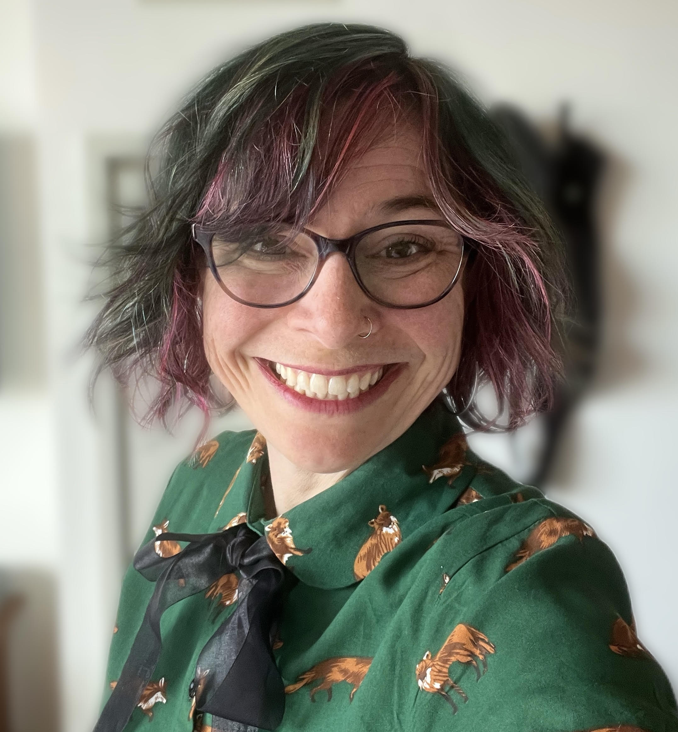 A person with glasses, blue and pink hair, smiling, wearing a green shirt with a tiger print denim jacket, and a black purse.