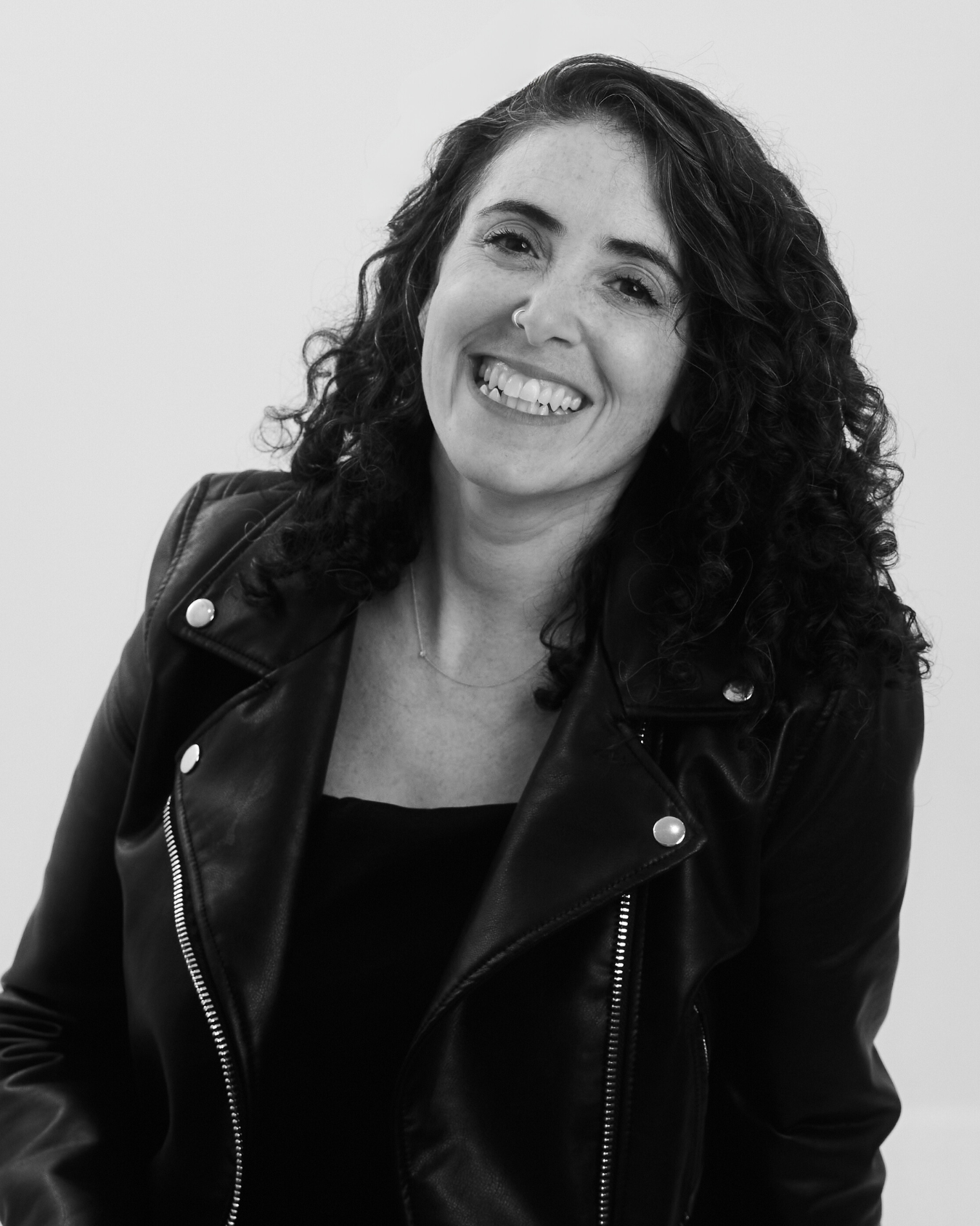 A smiling woman with curly hair wearing a leather jacket and a nose ring, captured in black and white.