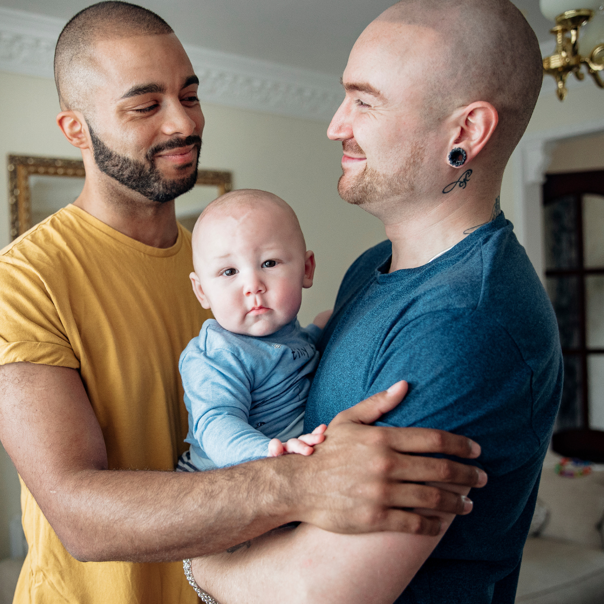 Two men, one with a beard and a shaved head, and the other with a bald head, holding a baby with a serious expression, inside a home with a mirror and chandelier in the background.
