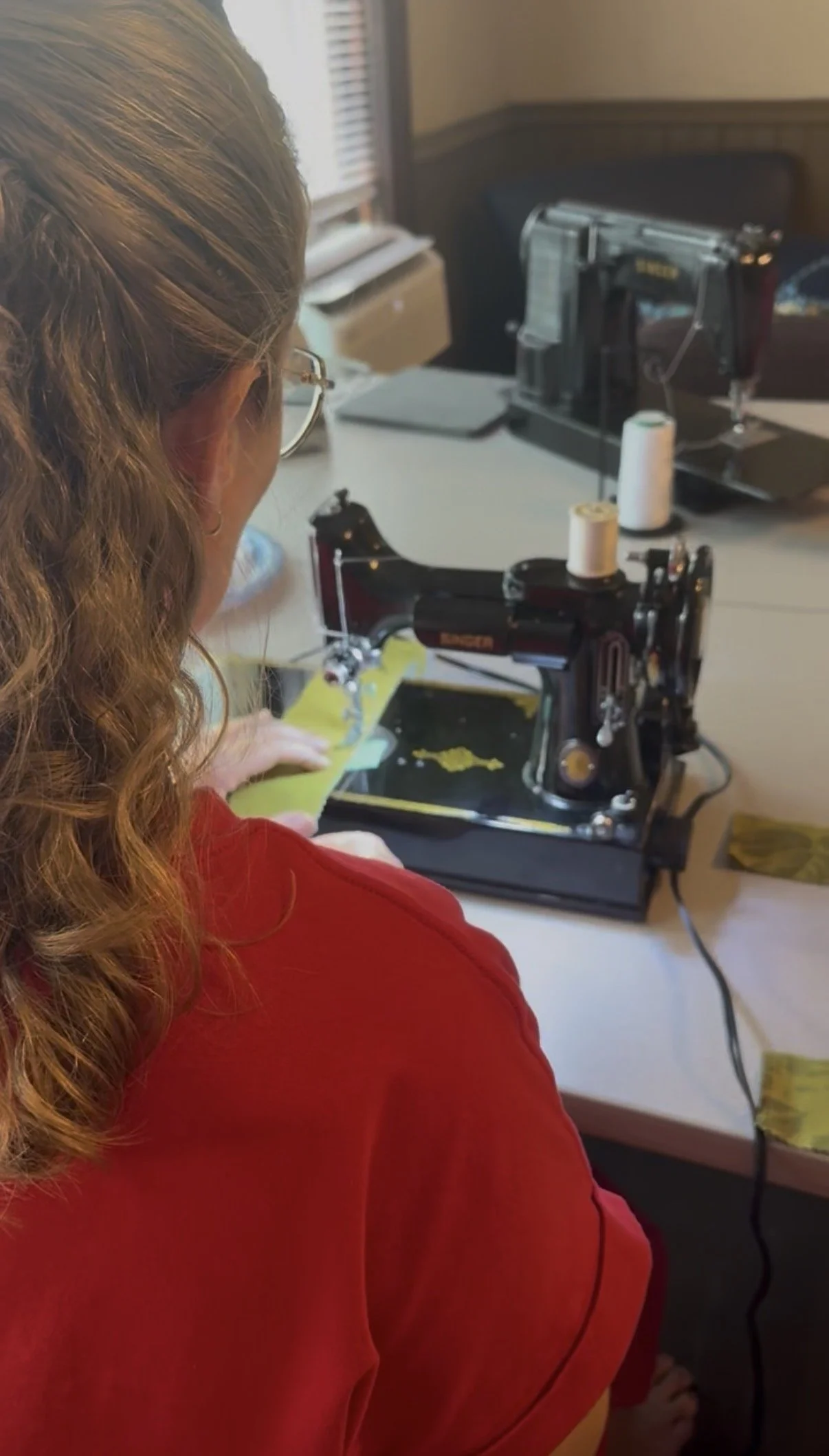 A person with long curly hair and glasses working on a sewing machine at a desk, with spools of thread and sewing materials around.