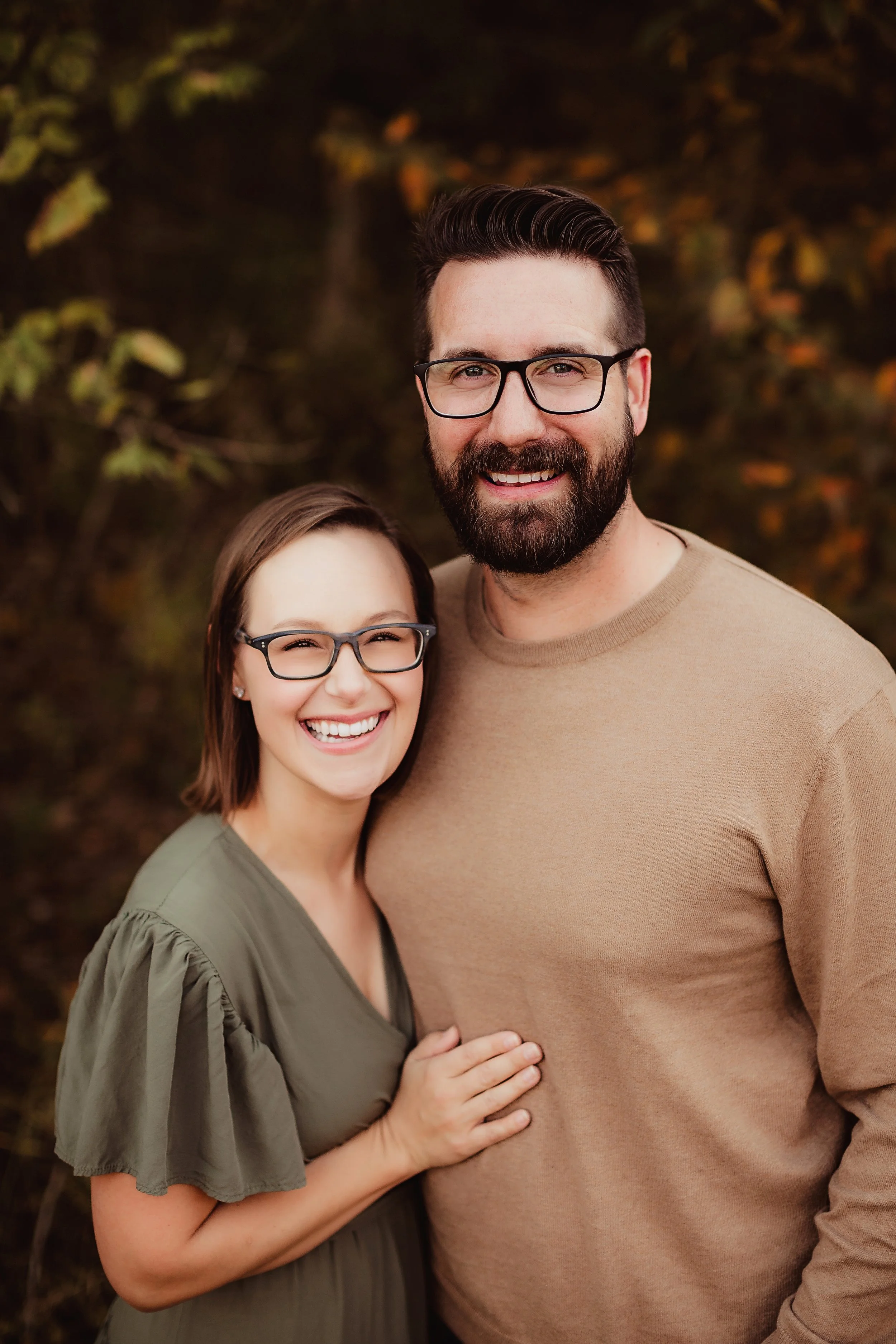 A smiling woman with glasses and shoulder-length brown hair, and a smiling man with glasses and a beard, standing outdoors in front of autumn foliage.