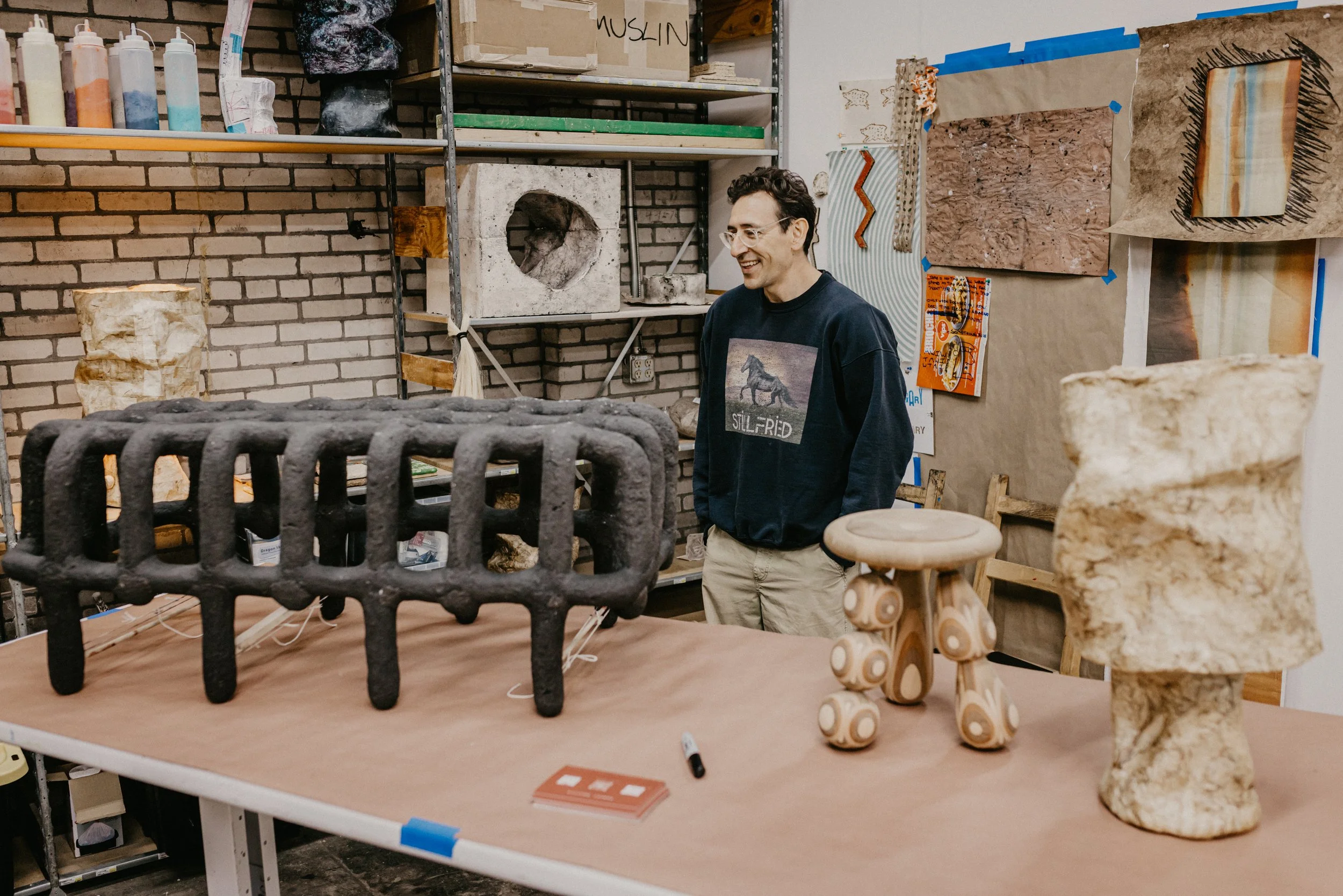 A person with glasses and a dark sweatshirt smiling in a workshop with various art pieces and sculptures around. The sculpture in the foreground resembles a black cast iron object, and there are other stone and mixed media artworks on the wall and table.