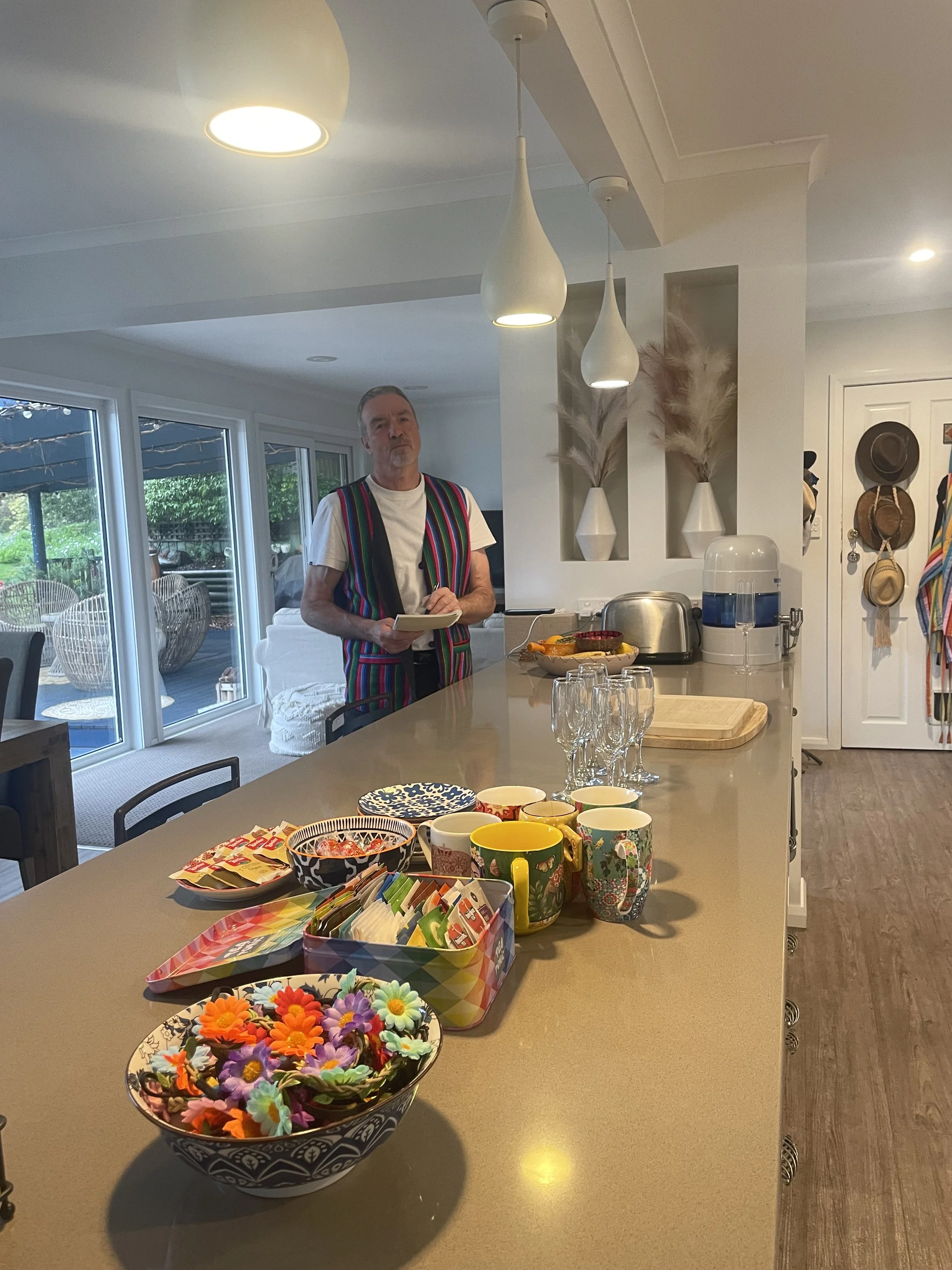 A man wearing a colorful striped vest stands in a modern kitchen, holding a notepad. The kitchen island is decorated with colorful cups, bowl of decorated flowers, and snacks. Large windows show an outdoor patio with chairs and greenery.