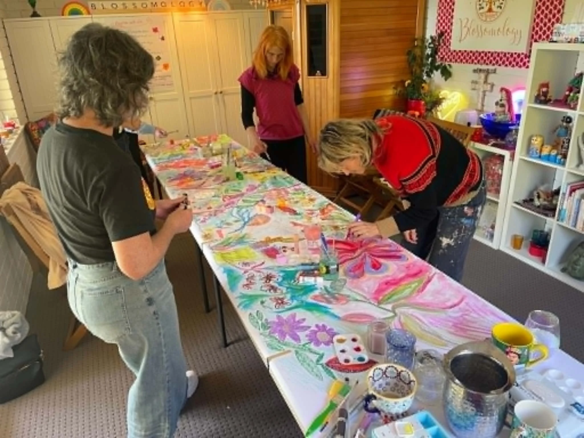 A group of people gathered around a table with colorful arts and crafts supplies in a cozy room decorated with rainbow and heart decorations.