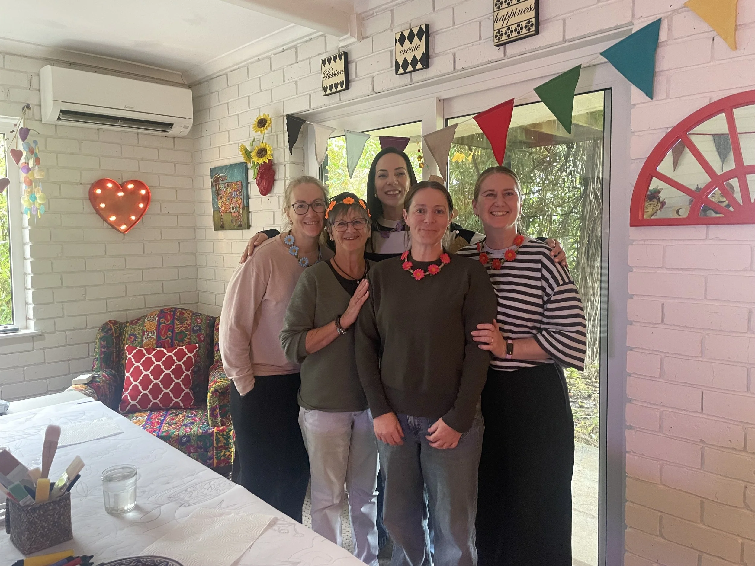 Five women standing indoors near a window, smiling and embracing each other. The room is decorated with colorful bunting, sunflower decorations, and a lit heart-shaped wall lamp. There are tables with white tablecloths and a window showing green foli