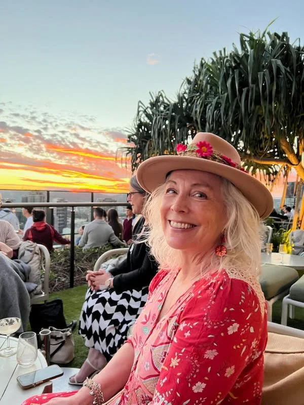 A woman with blonde hair, wearing a pink floral dress and a straw hat decorated with pink flowers, smiling at sunset on a rooftop.