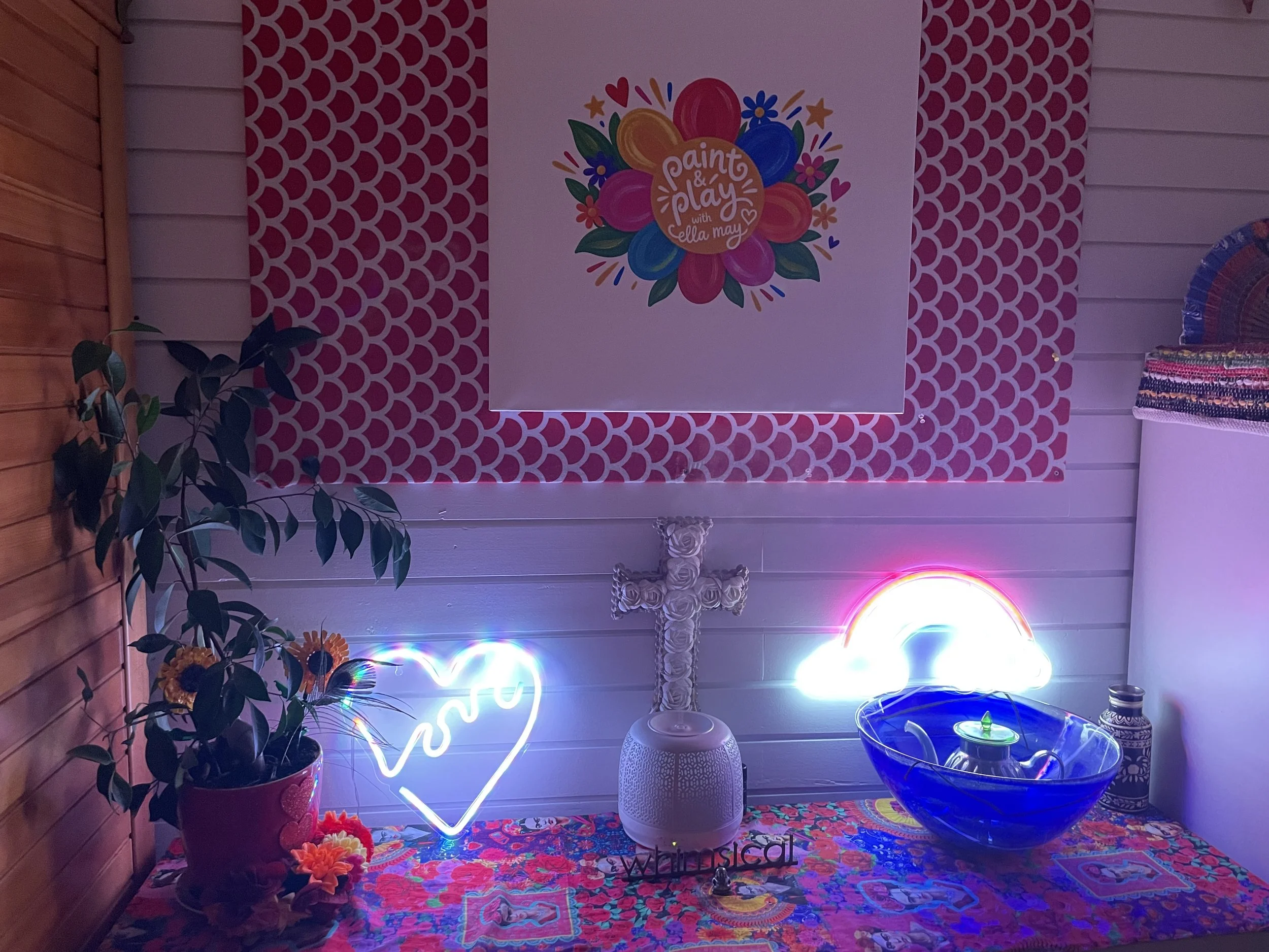 A colorful decorative setup featuring a painted pink and red backdrop with a large floral sign that reads 'Paint & Play with Ella May', a neon heart-shaped light, a potted plant, a white textured table lamp, a vibrant tablecloth, a large blue glass b
