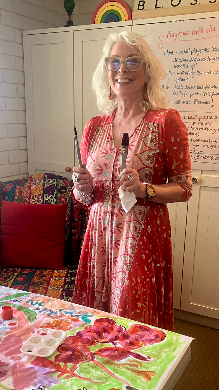 A woman with blonde curly hair and glasses smiling, wearing a red floral dress, standing in a room with colorful decorations, holding painting tools in both hands, with a painting and art supplies on the table in front of her.