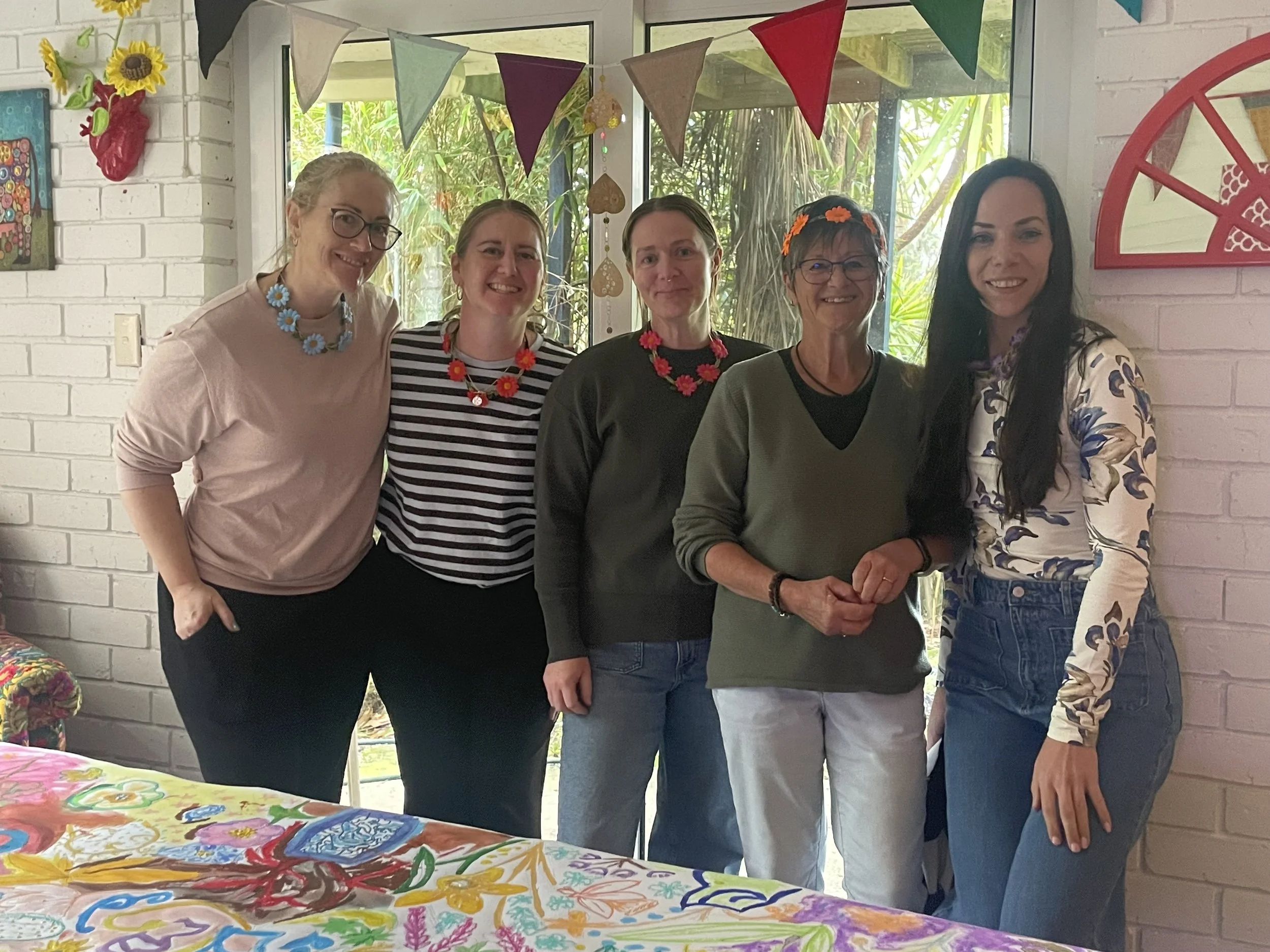 Five women standing together indoors in front of a glass window with greenery outside. They are smiling and wearing colorful necklaces and casual clothes. The room has decorations including bunting and framed artwork, with a table covered in a bright