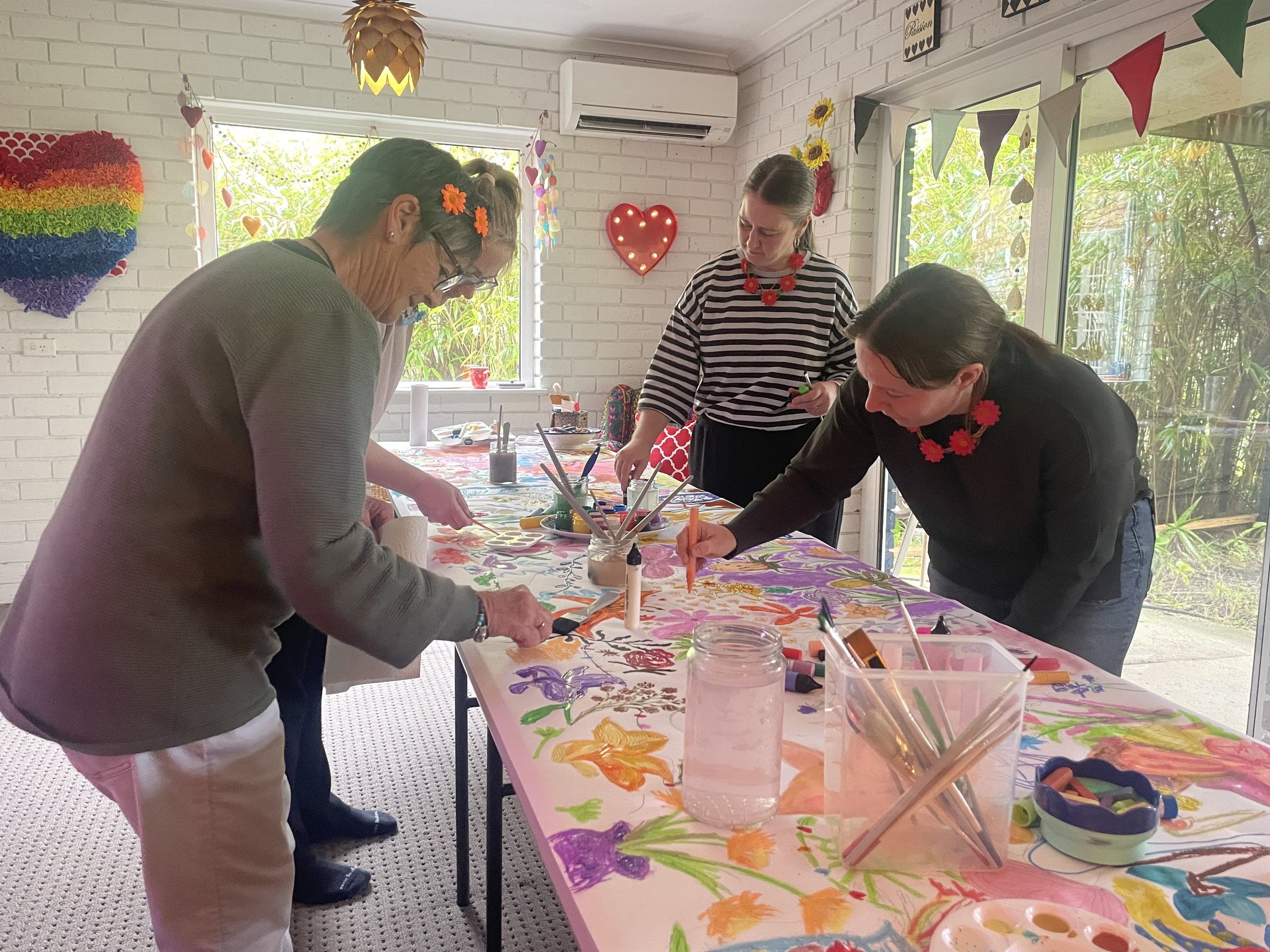 People participating in a group painting activity around a table covered with colorful artwork, inside a room decorated with heart and bunting decorations, with large windows showing green outdoor scenery.