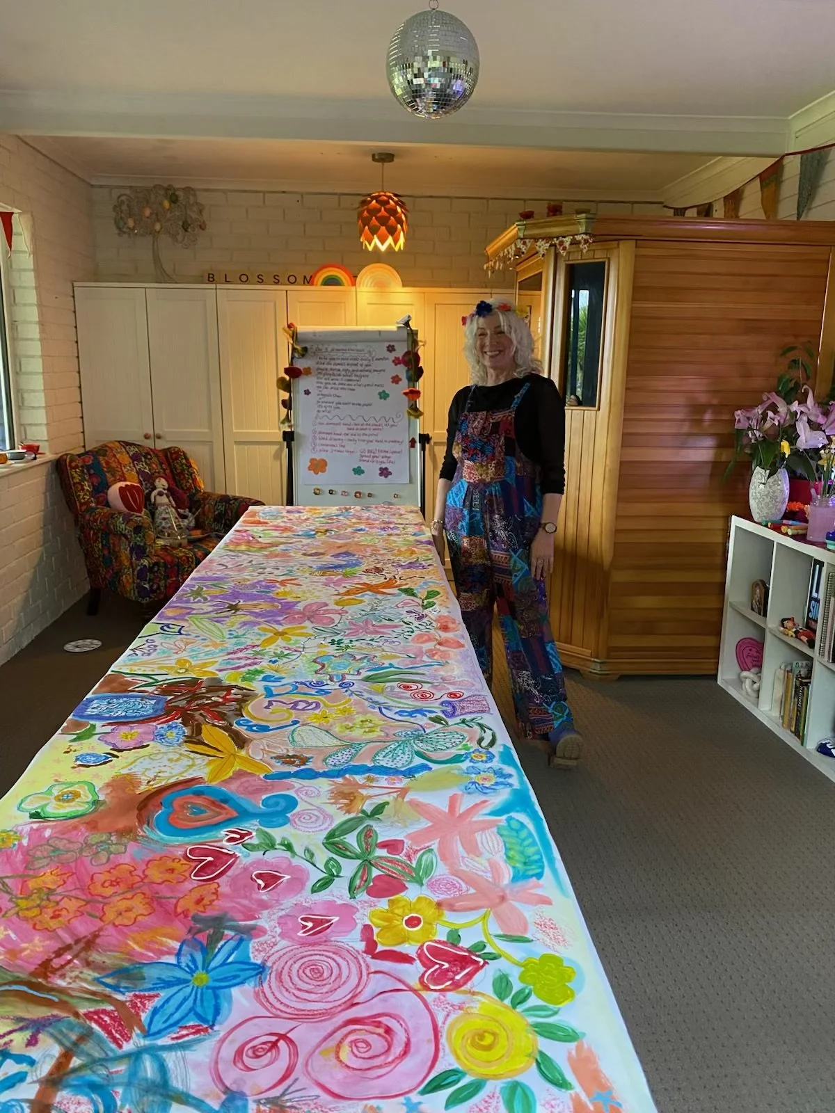 A woman with curly gray hair smiling at a colorful, painted table in a cozy room. The room has decorated walls, a white cabinet, a white bookshelf with books, pink flowers in a vase, and a disco ball hanging from the ceiling.