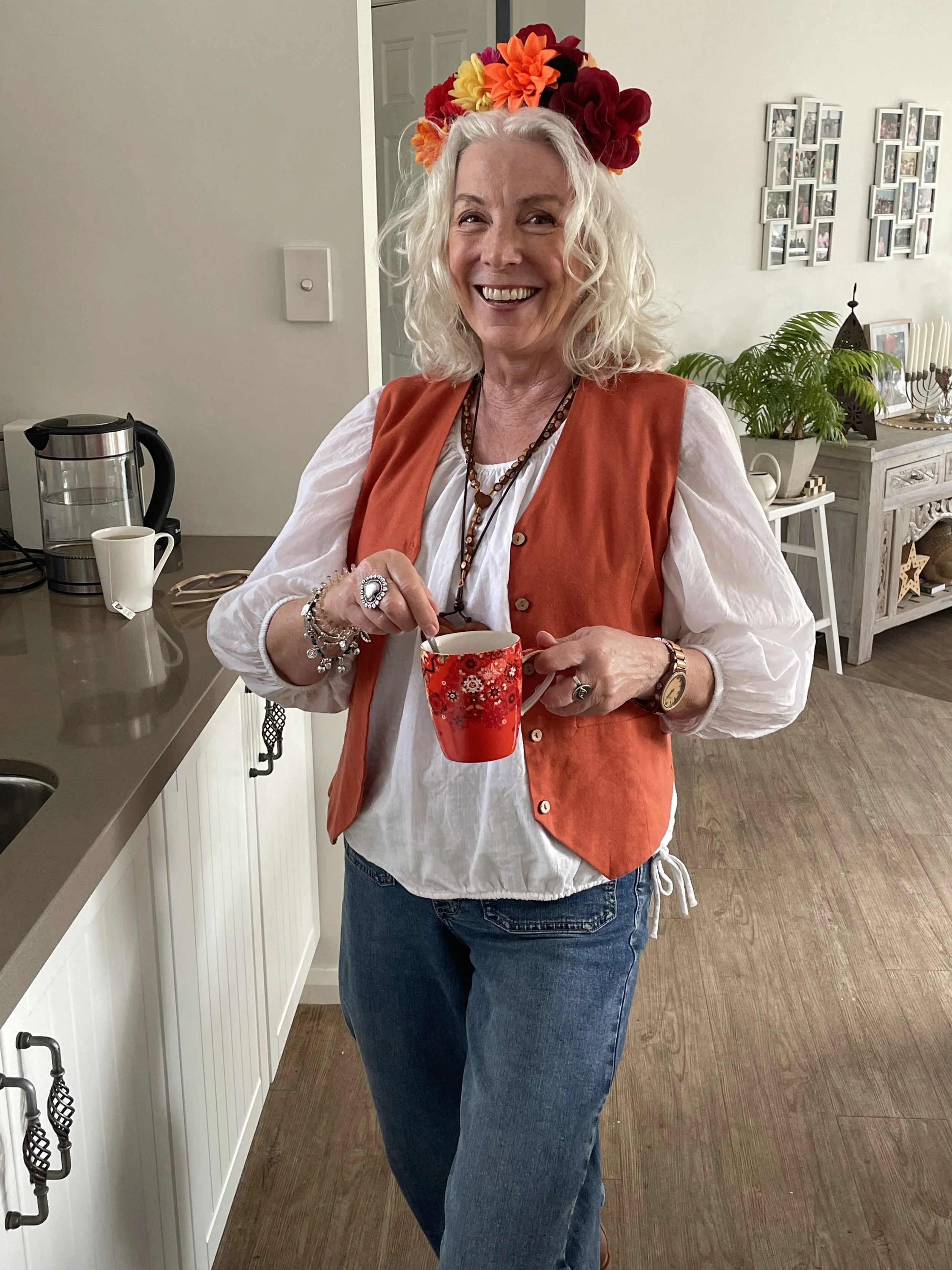 A woman with curly blonde hair wearing a floral headband, orange vest, and jewelry, holding a red floral mug while smiling in a cozy room with a white console table, green potted plant, and framed photos on the wall.