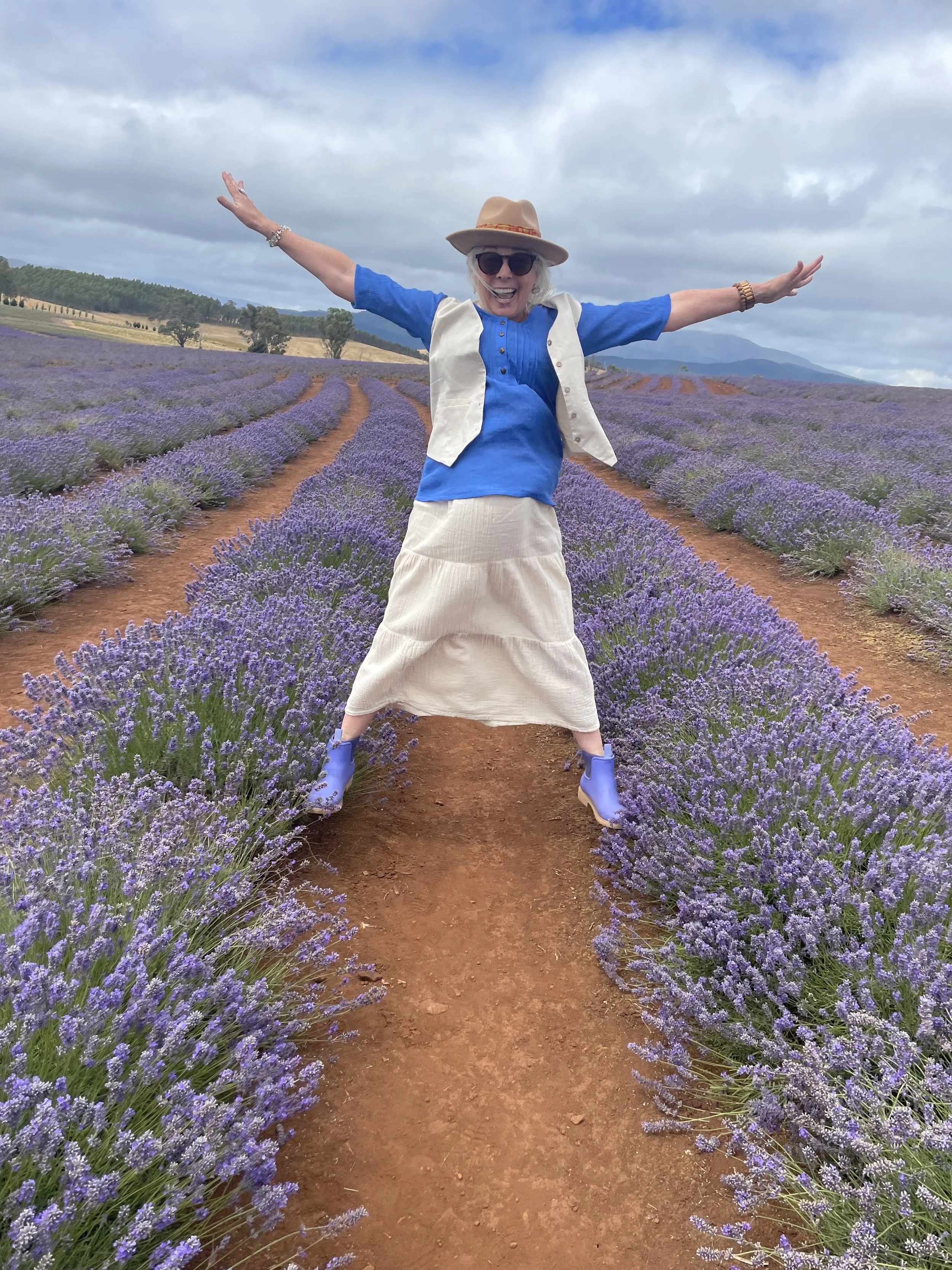 An elderly woman standing in a lavender field with arms outstretched, wearing a beige hat, sunglasses, a blue top, a white vest, a cream-colored skirt, and blue boots, smiling.