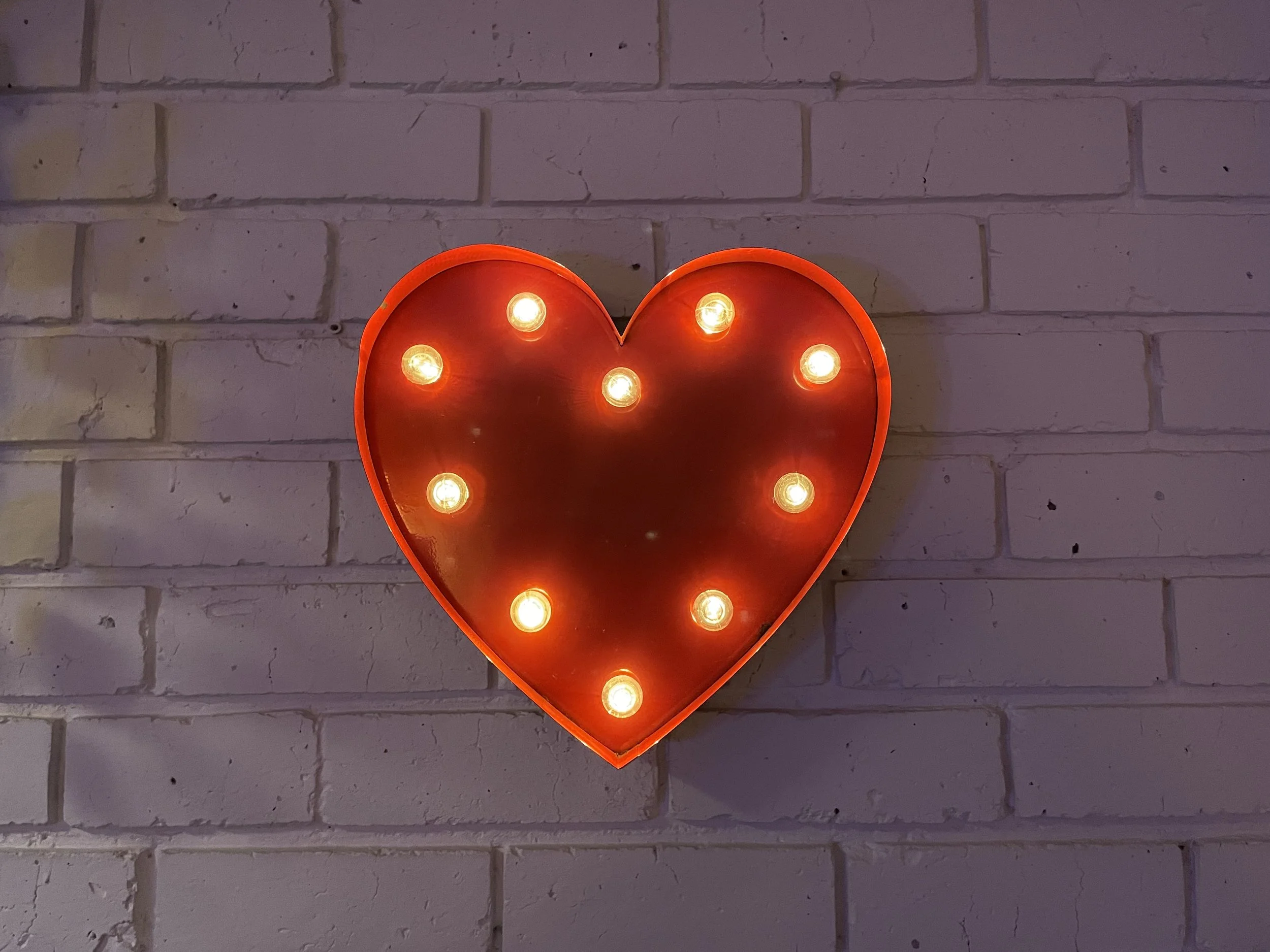 Red heart-shaped light fixture with exposed light bulbs mounted on a brick wall.