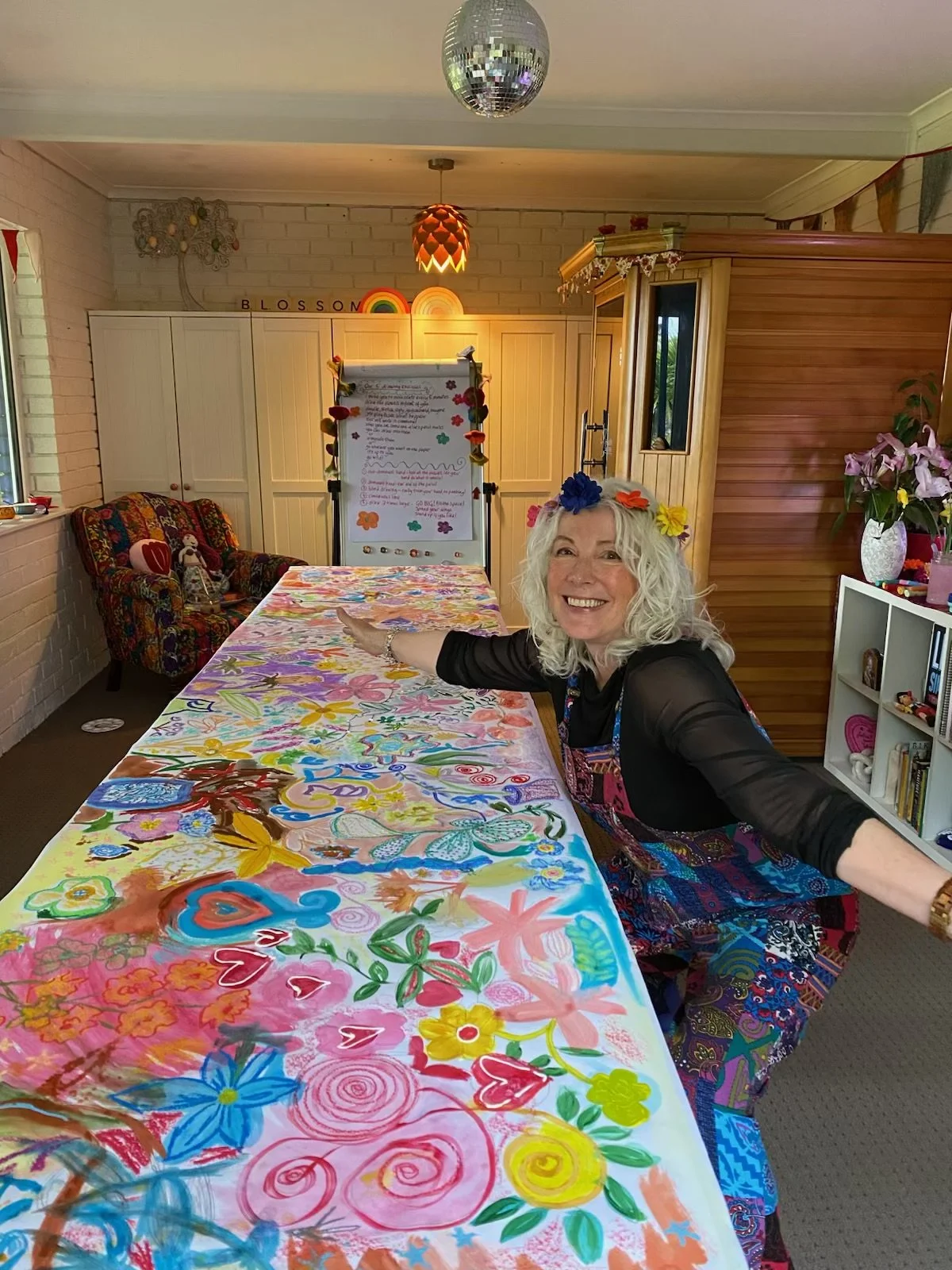 A smiling woman with curly gray hair and a colorful flower headband leans on a table covered with vibrant, colorful artwork. The room has a white brick wall, a couch with floral upholstery, and a whiteboard with notes. There are also decorative items