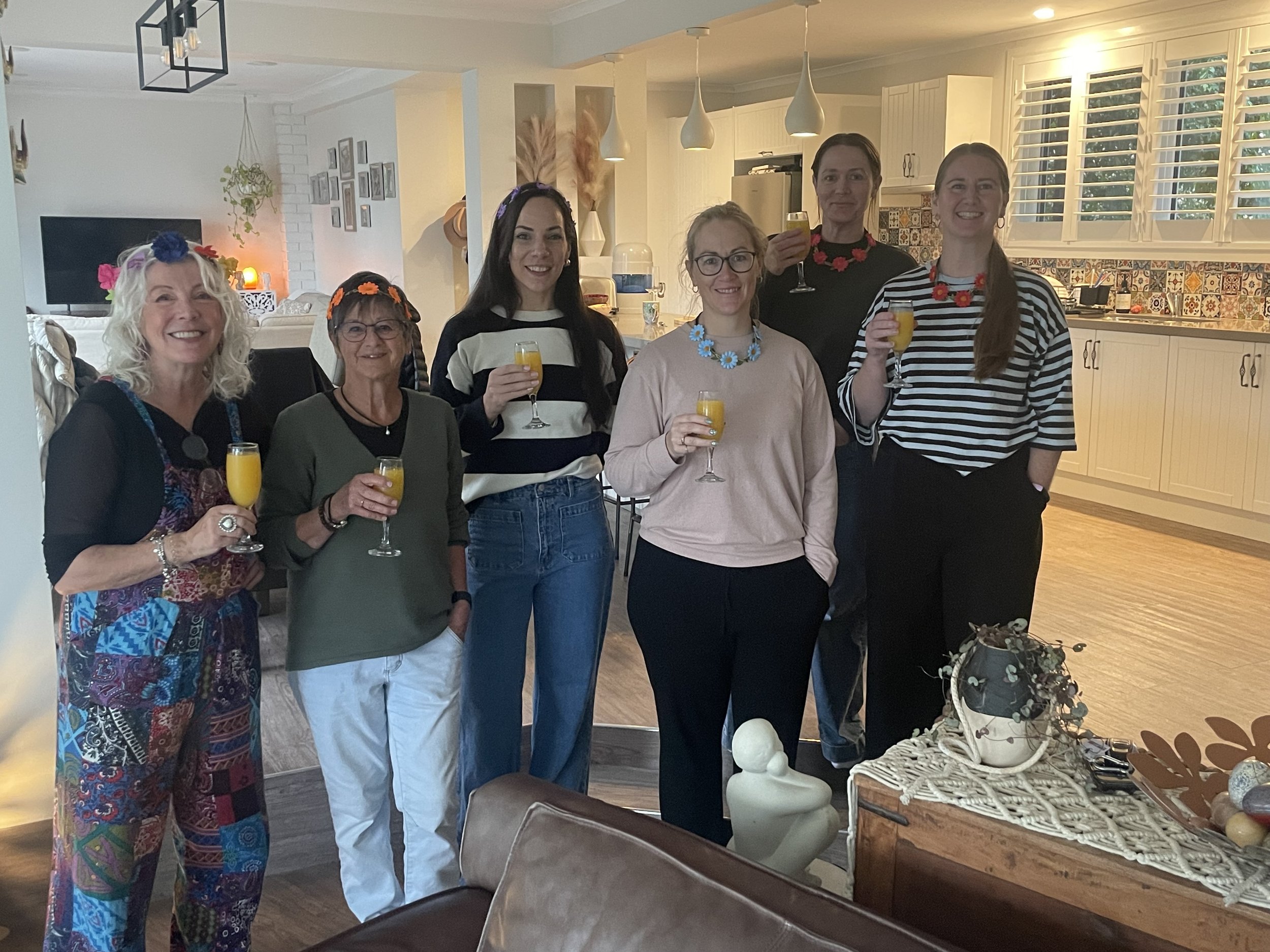 A group of seven women standing together inside a modern kitchen, smiling and holding glasses of orange juice, celebrating an occasion.